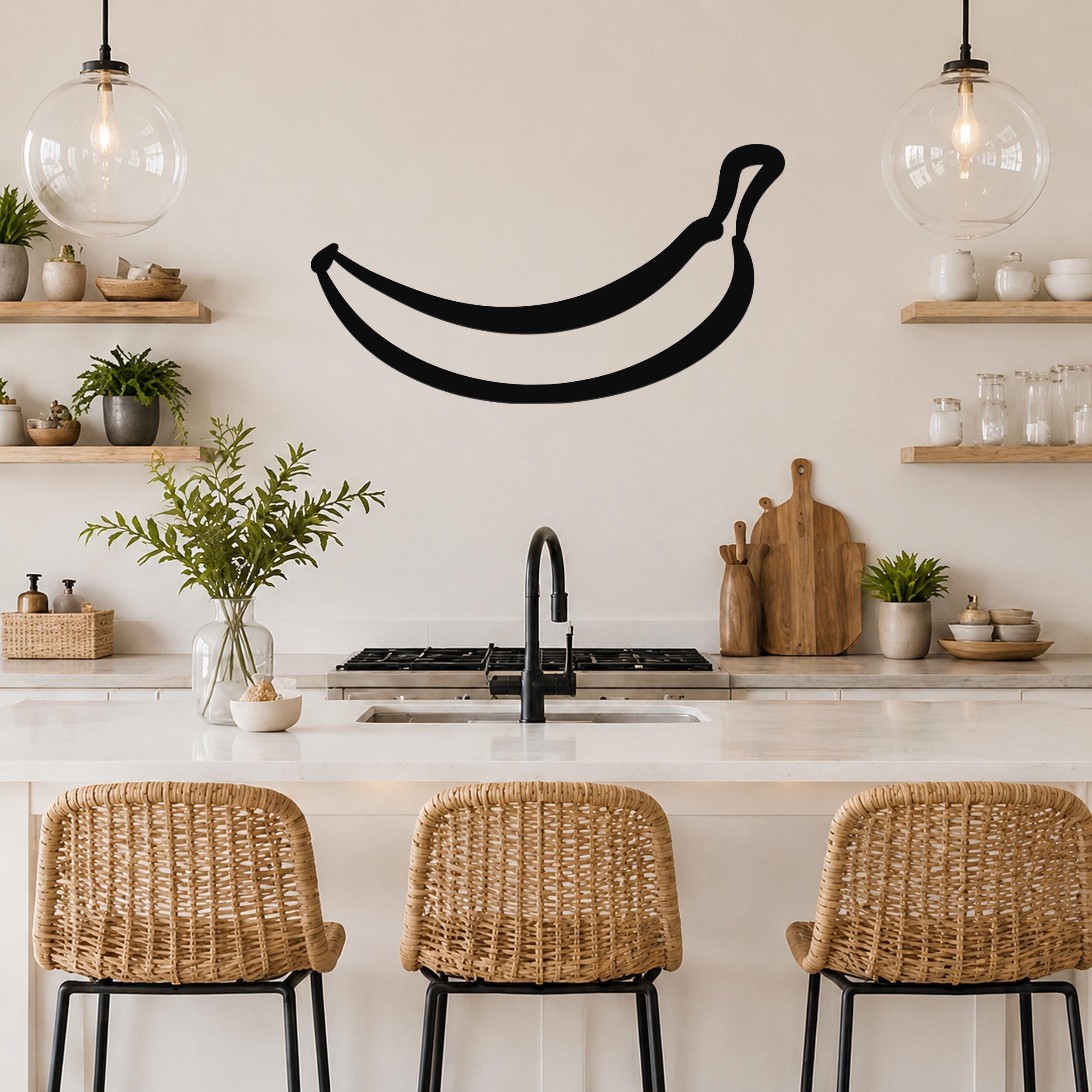 Black metal banana wall art silhouette mounted above a kitchen counter, displayed in a modern kitchen with wooden shelves and pendant lights.
