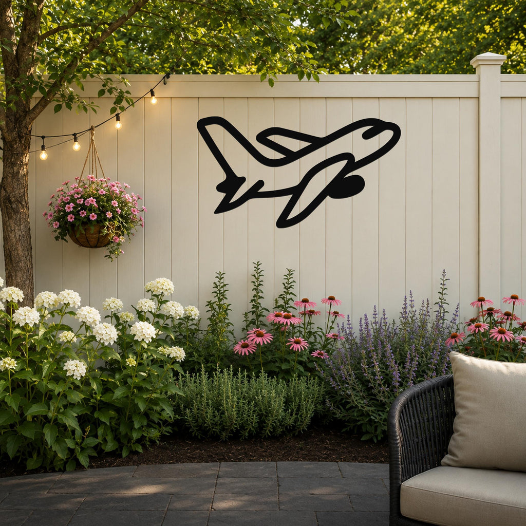 Black metal airplane wall art silhouette mounted on a white outdoor fence, above a garden bed with flowers and string lights.