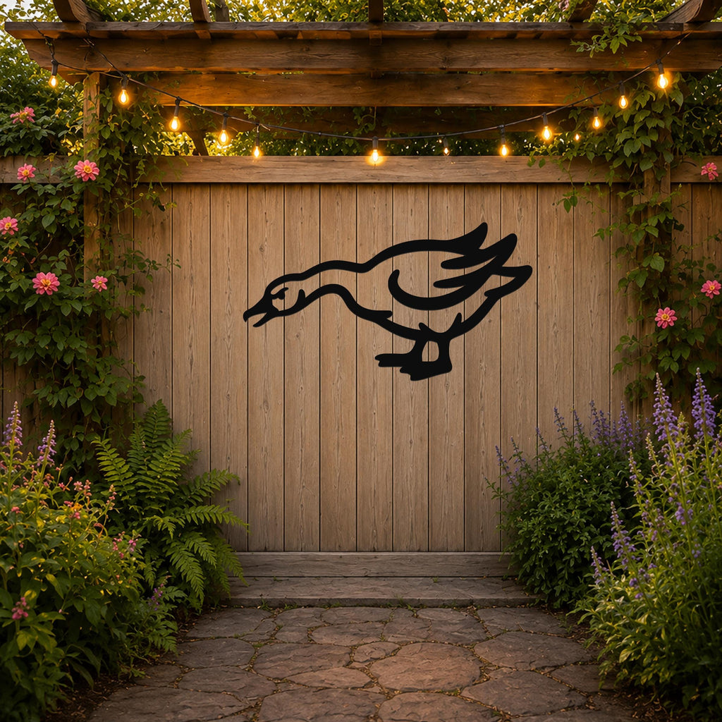 Black metal goose wall art with a silhouette cutout design, mounted on a wooden fence in a garden setting with string lights and flowering plants.