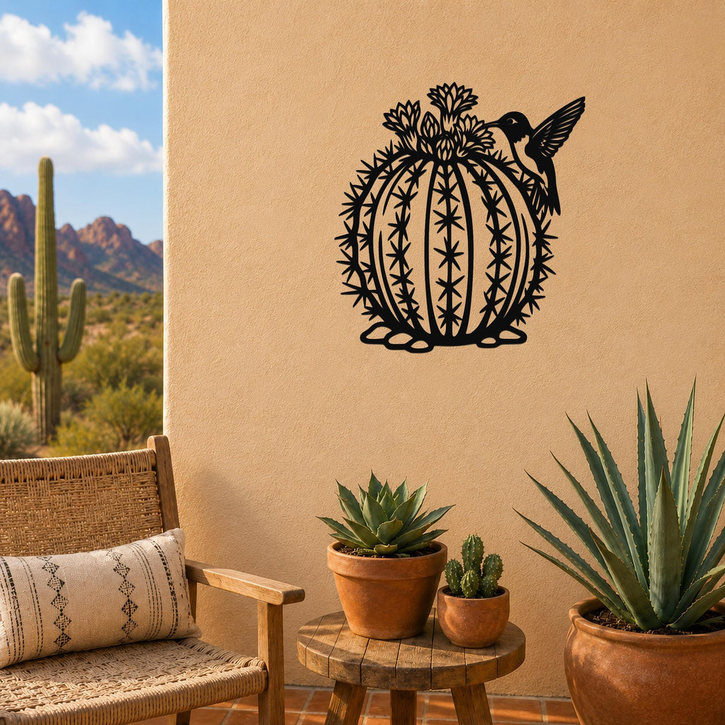 Black metal barrel cactus wall art with hummingbird and flower cutouts, mounted on a tan outdoor wall above a small wooden table with potted succulents.