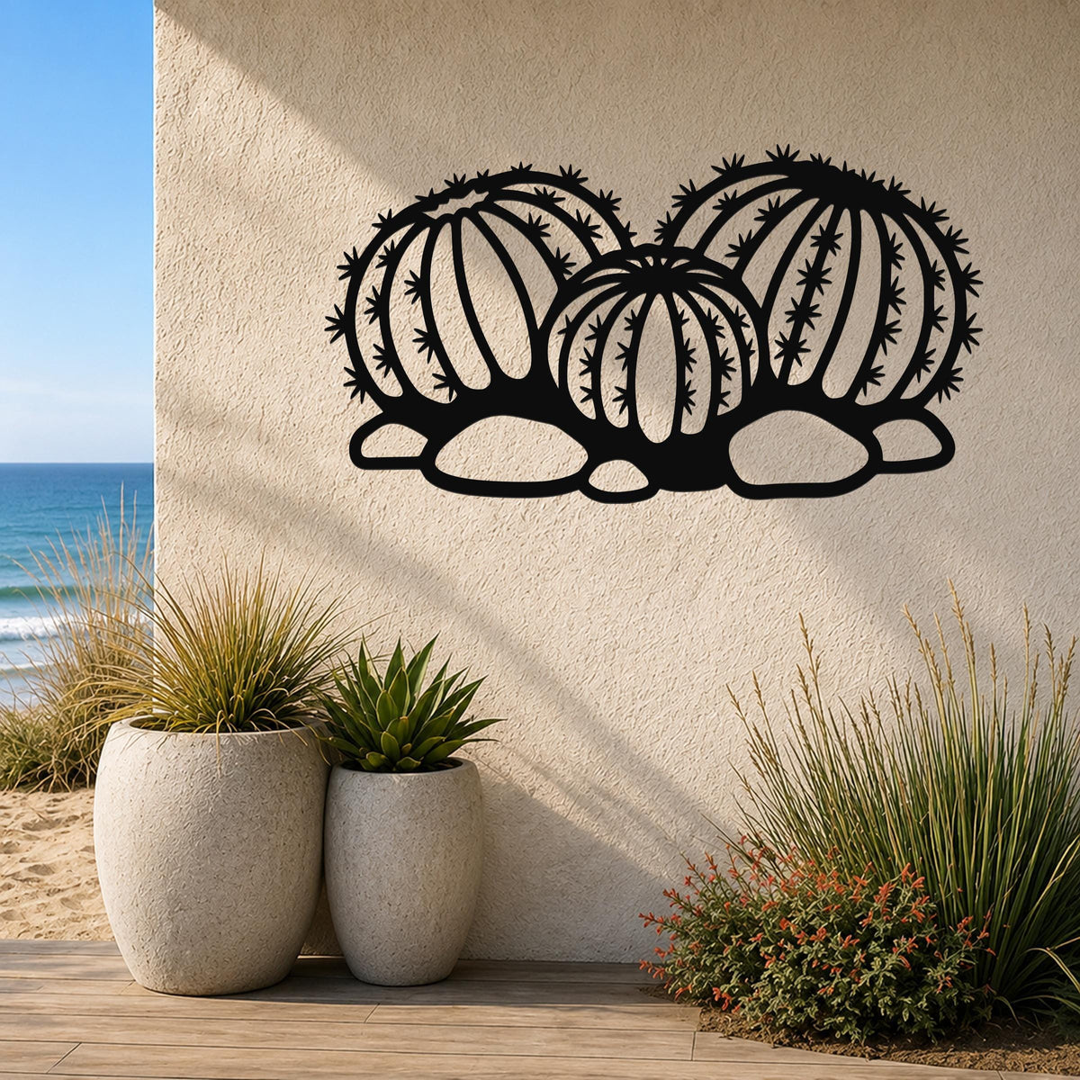 Black metal barrel cactus wall art with spines and segmented cutouts, mounted on an outdoor wall above potted plants near a beach.