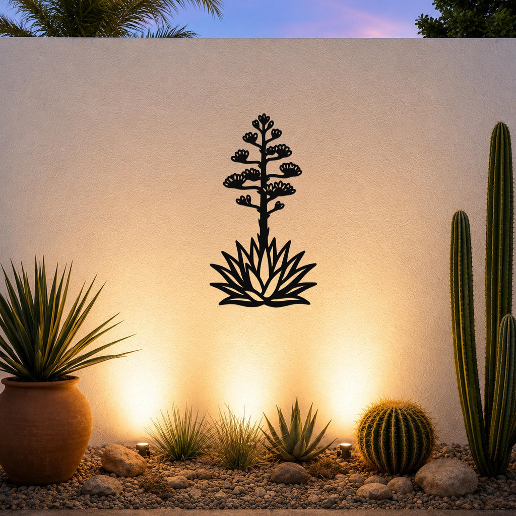 Black metal agave wall sculpture with blooming flower and spiky leaves cutout, mounted on a light-colored outdoor wall above a desert garden with cacti and warm ground lighting.
