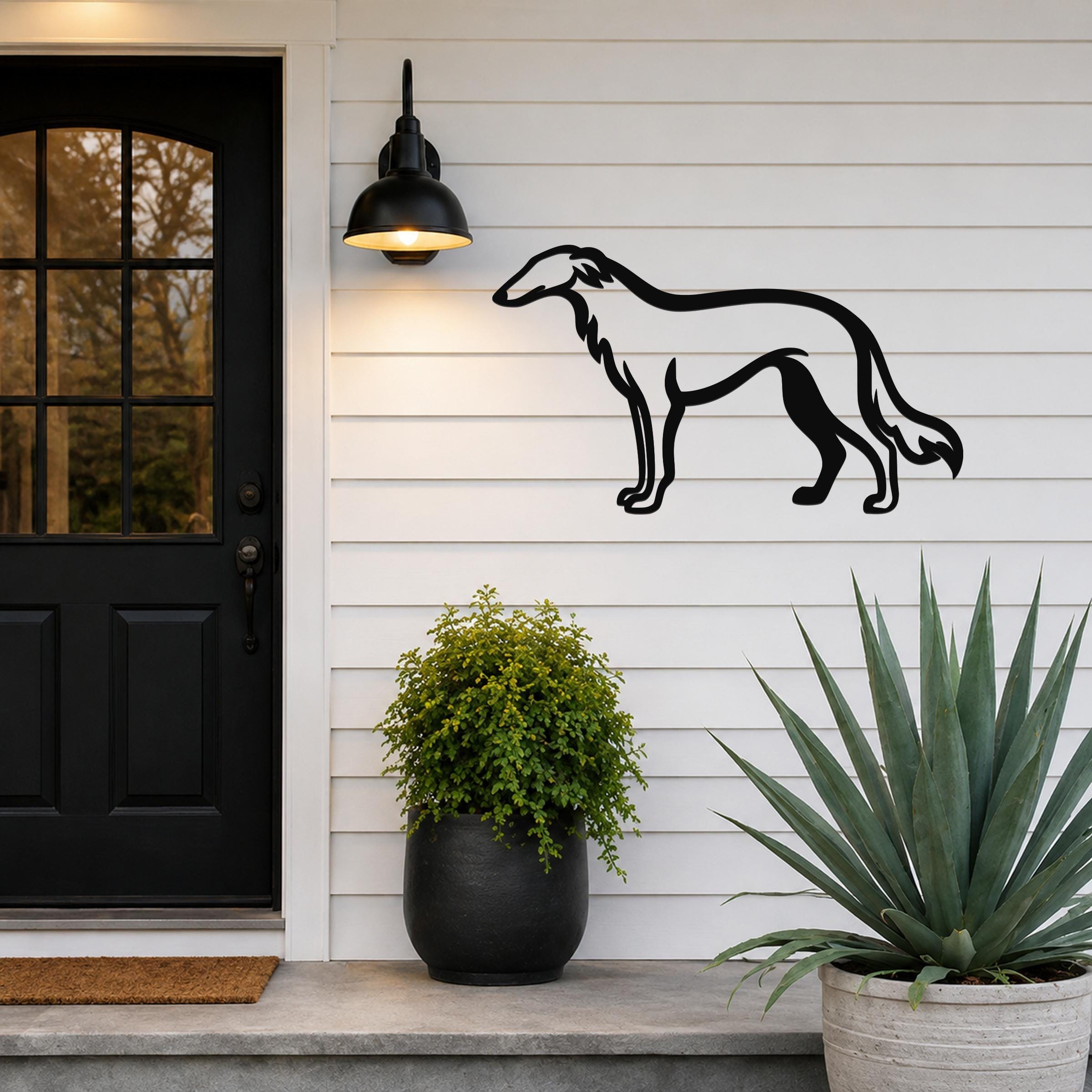 Black metal Borzoi dog wall art silhouette mounted on a white siding wall beside a black front door, with a lit outdoor sconce above and potted plants below.