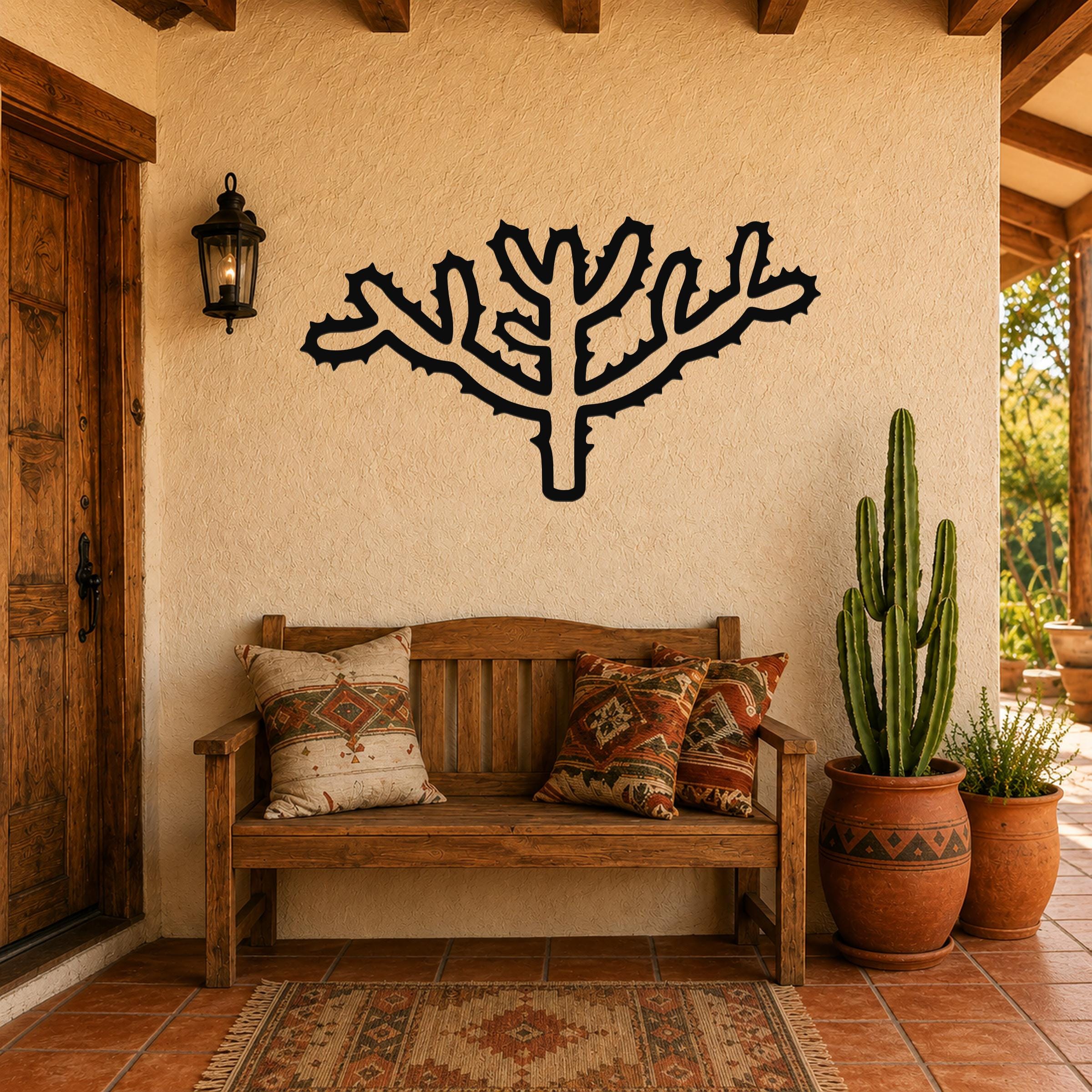 Black metal wall art in the silhouette of a cholla cactus with branching arms and spines, mounted on a stucco wall above a wooden bench in a patio entryway.