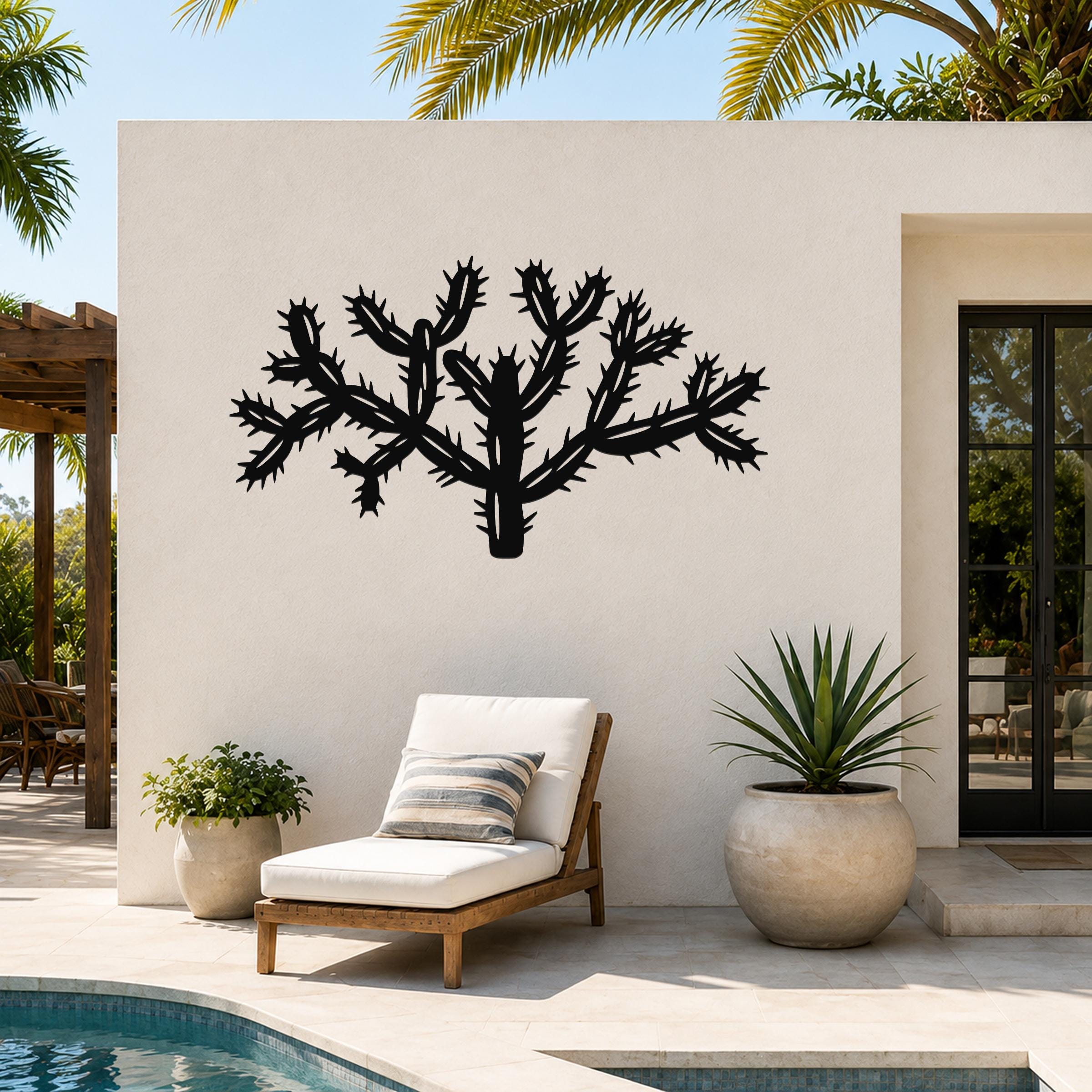Black metal Cholla cactus wall art with textured spine cutouts, mounted on an outdoor patio wall above a lounge chair.