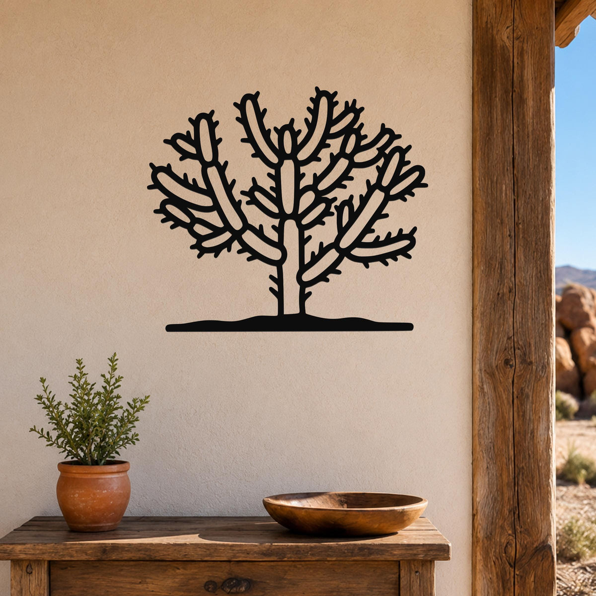 Black metal Cholla cactus wall art with detailed branch cutouts, mounted on a beige wall above a wooden table in a rustic outdoor living space.