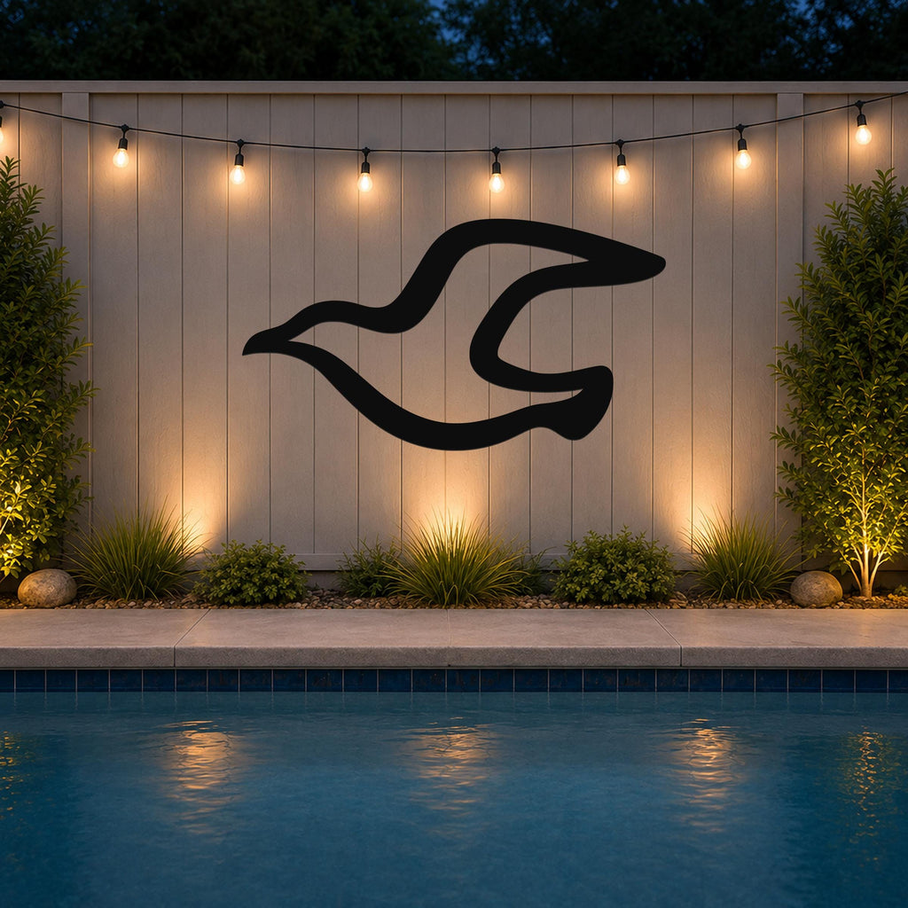 Black metal seagull wall art with flowing wing silhouette, mounted on a light-colored outdoor wall above a pool at dusk.