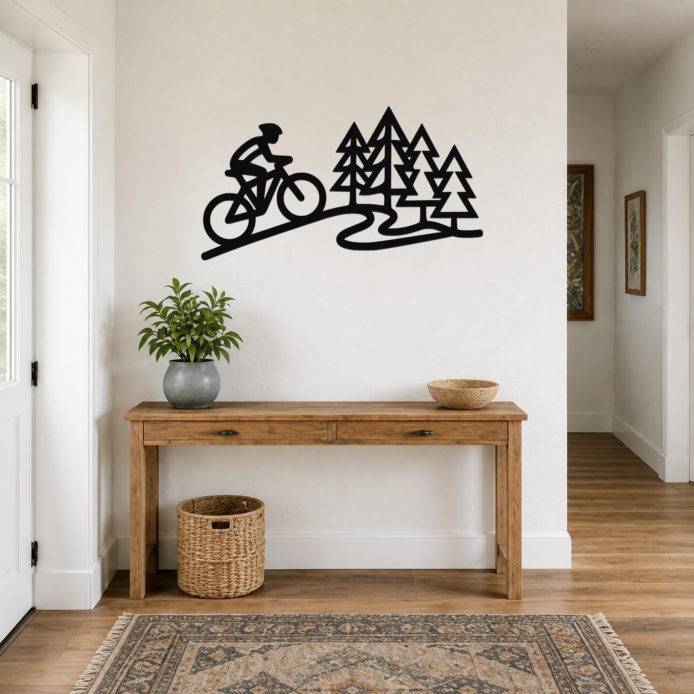 Black metal wall art silhouette of a cyclist on a trail with pine trees, mounted above a wooden console table in a hallway.