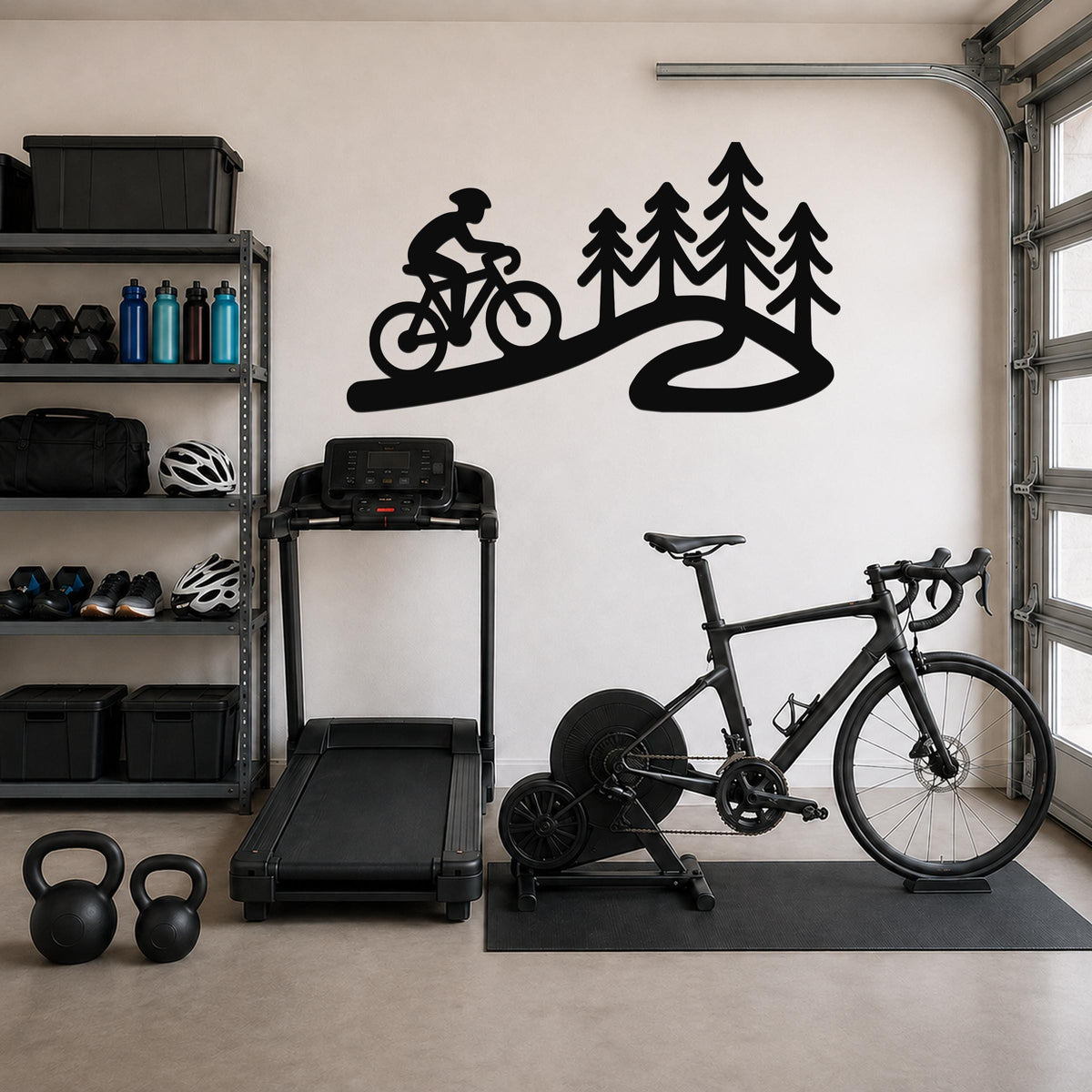 Black metal wall art silhouette of a cyclist on a winding path with pine trees, mounted on a garage wall above a treadmill and a bike on a trainer.