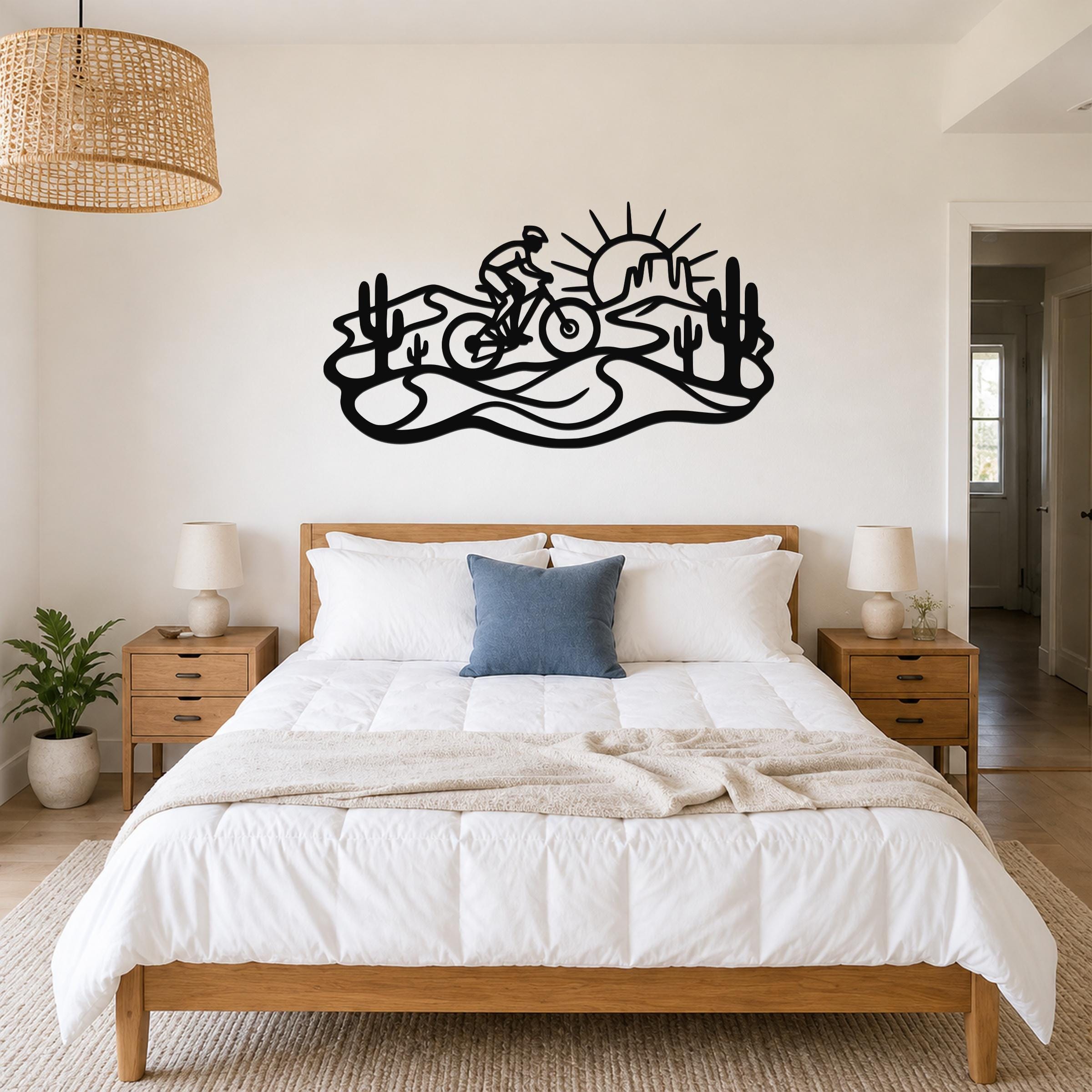 Black metal wall art silhouette of a cyclist on a desert trail with cacti and a sun, mounted above a bed in a bedroom.