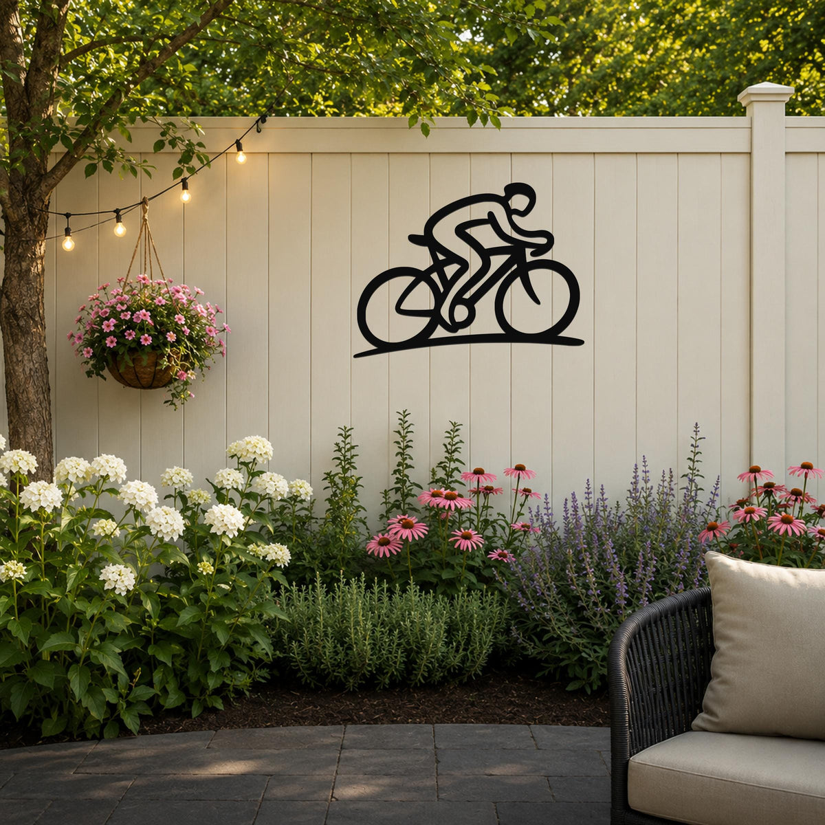 Black metal wall art of a cyclist silhouette, mounted on a light-colored outdoor fence above a flower bed with string lights and a hanging basket.