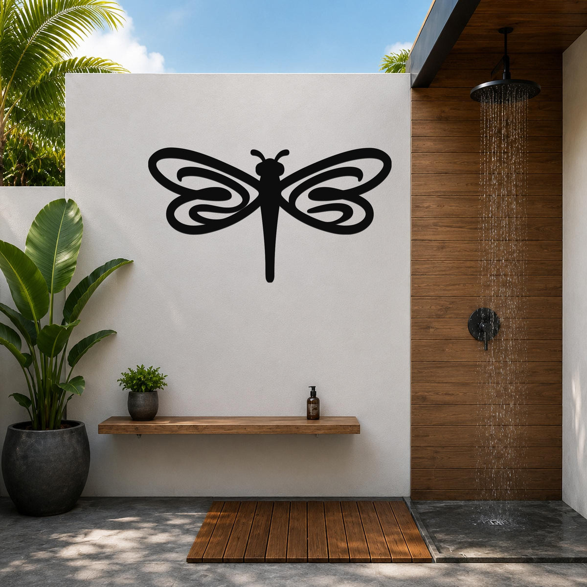 Black metal dragonfly wall art with intricate wing cutouts, mounted on a white outdoor wall above a wooden shelf in a patio shower area.