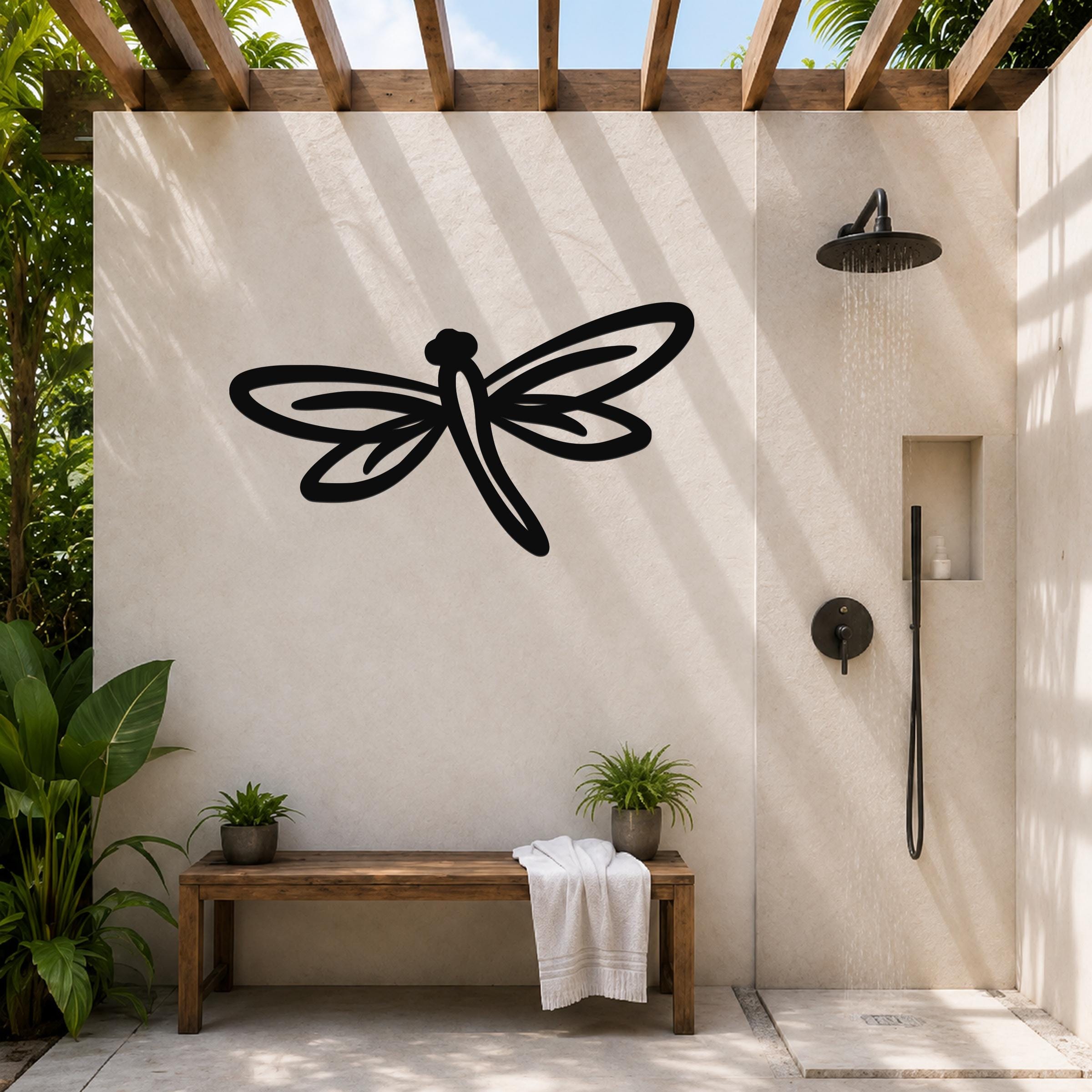 Black metal dragonfly wall art with cutout wings and body, mounted on a beige outdoor shower wall above a wooden bench.