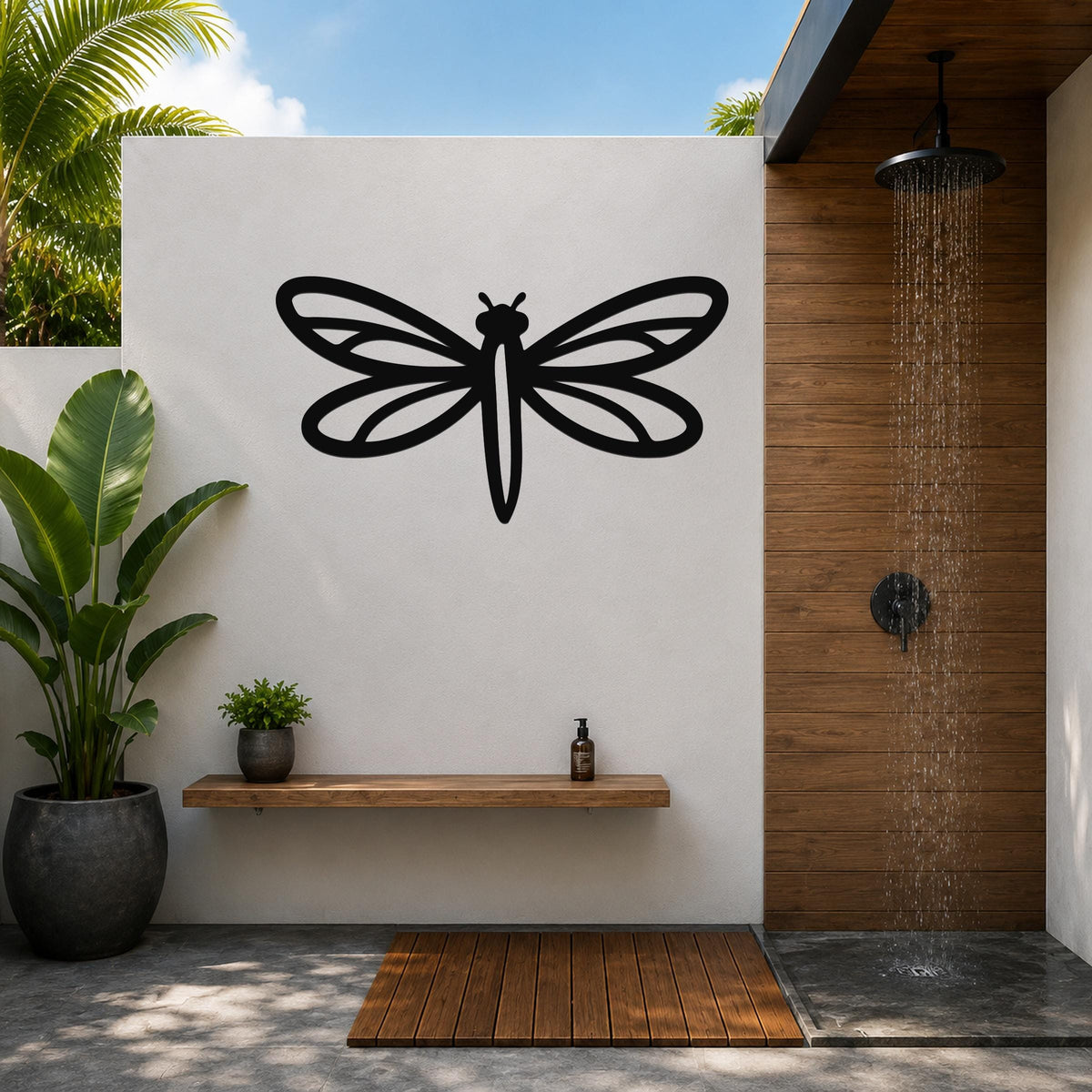Black metal dragonfly wall art with detailed wing cutouts, mounted on a white outdoor wall above a wooden shelf in a modern bathroom.