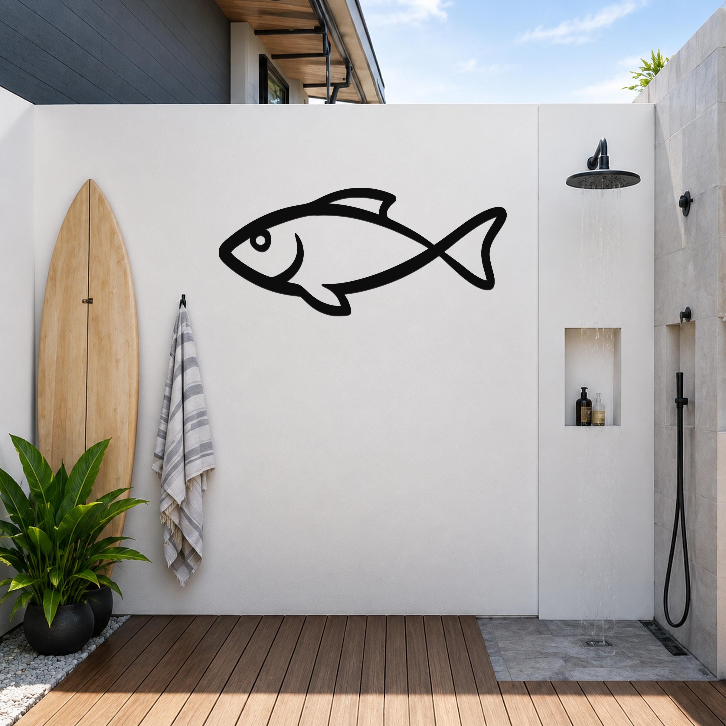 Black metal fish silhouette wall art mounted on a white outdoor wall beside a shower, with a surfboard and towel nearby.