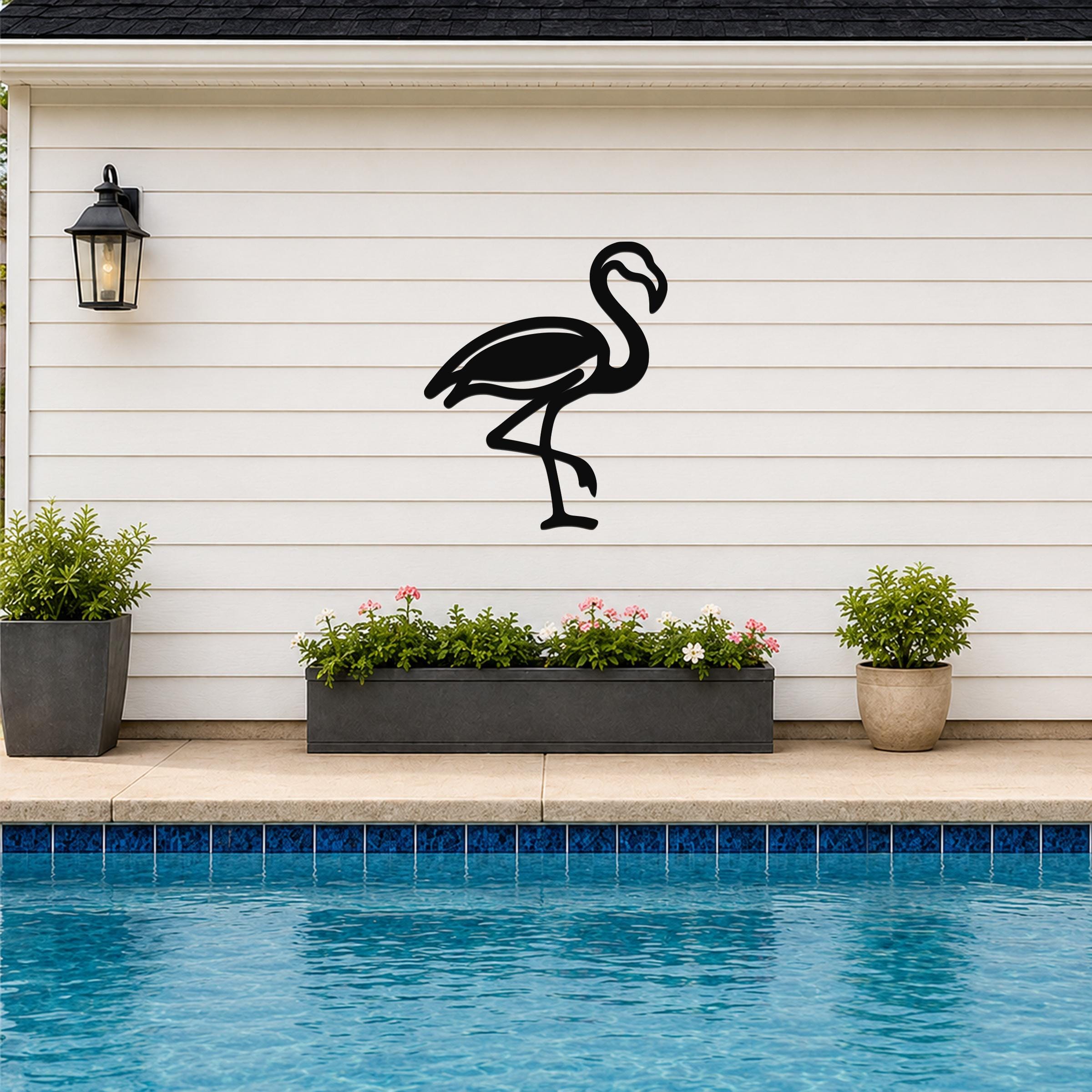 Black metal flamingo wall art silhouette mounted on a white horizontal siding wall above a poolside planter with flowers and plants.