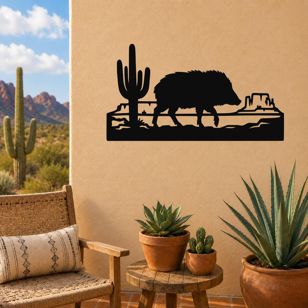 Black metal wall art featuring a javelina silhouette walking past a saguaro cactus and canyon rock formation, mounted on a tan outdoor wall above a small wooden table with potted succulents.