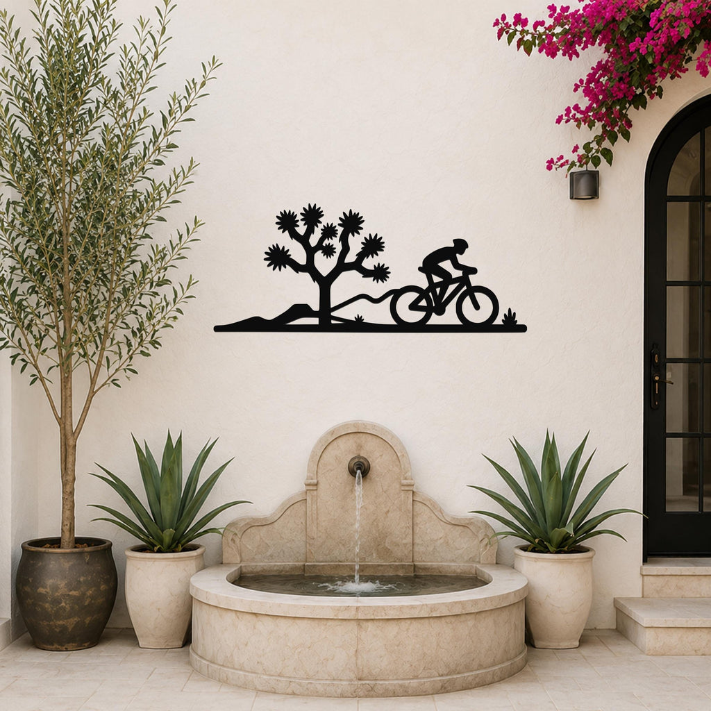 Black metal wall art silhouette of a cyclist and Joshua tree, mounted on a white outdoor wall above a stone fountain.
