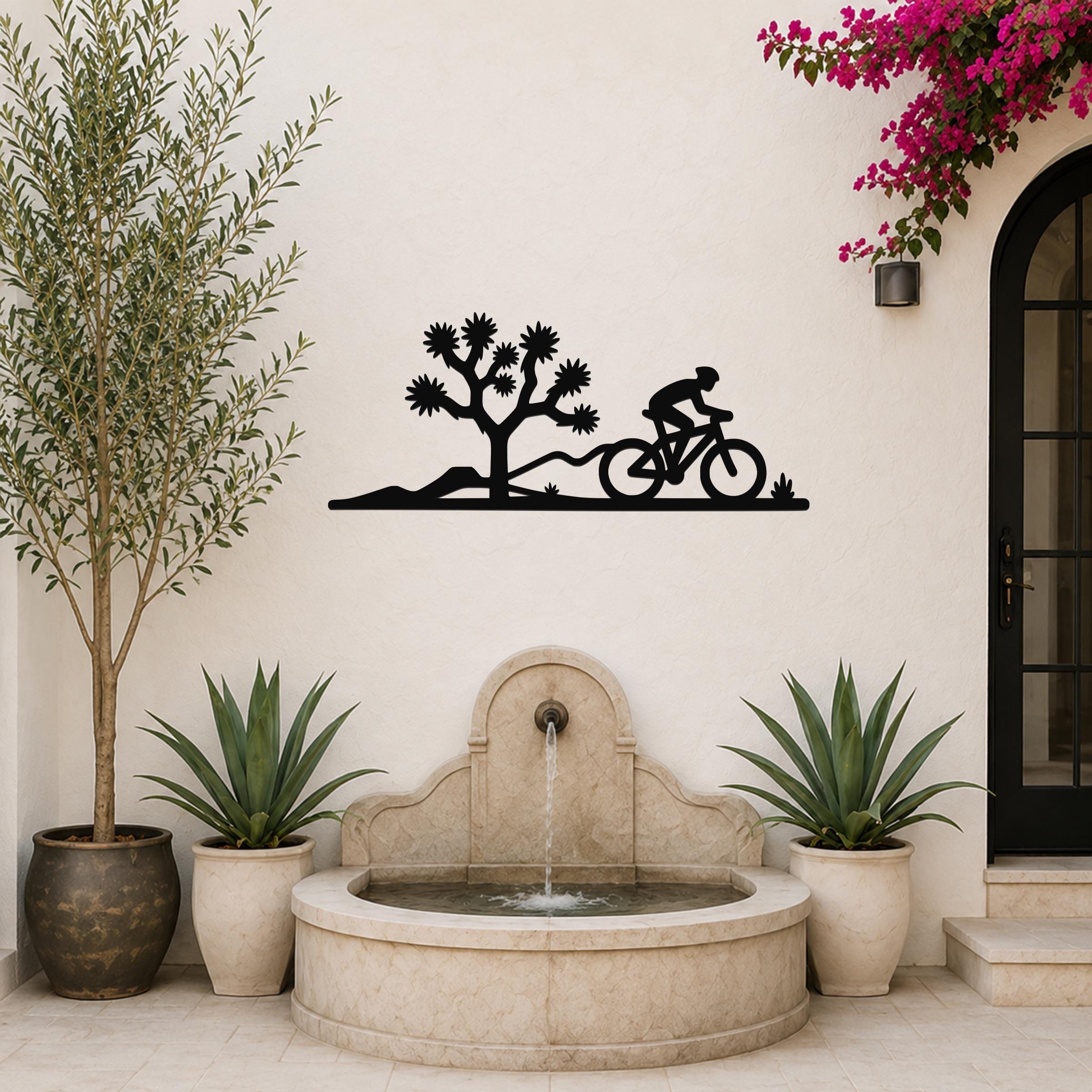 Black metal wall art silhouette of a cyclist and Joshua tree, mounted on a white outdoor wall above a stone fountain.