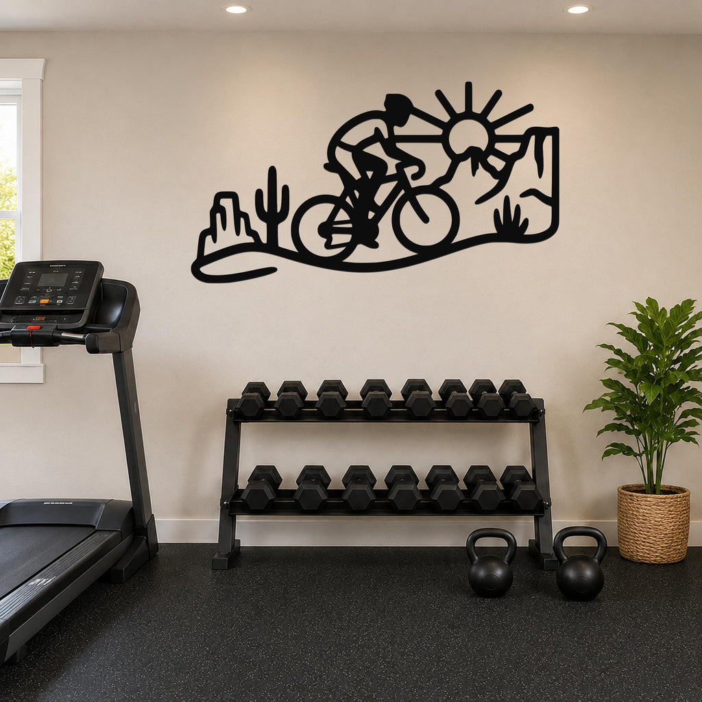 Black metal wall art silhouette of a cyclist on a desert trail with cacti and a sun, mounted on a wall above a dumbbell rack in a home gym.