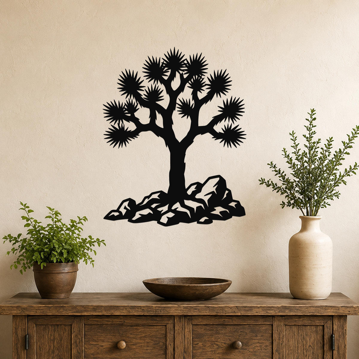 Black metal wall art silhouette of a Joshua tree with spiky branches and rocky base, mounted on a beige wall above a wooden console table.