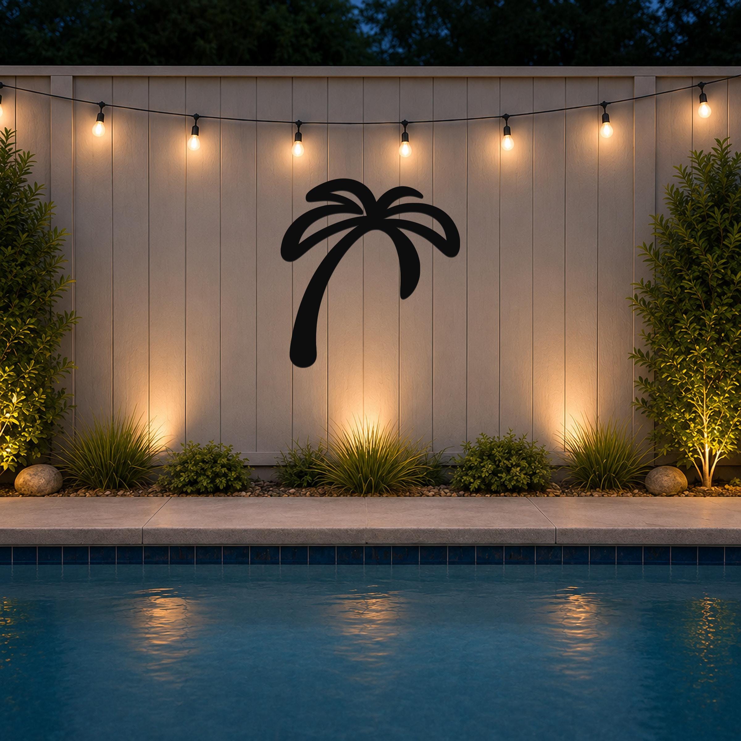Black metal palm tree wall art silhouette mounted on a light-colored outdoor wall above a pool, illuminated by string lights at dusk.