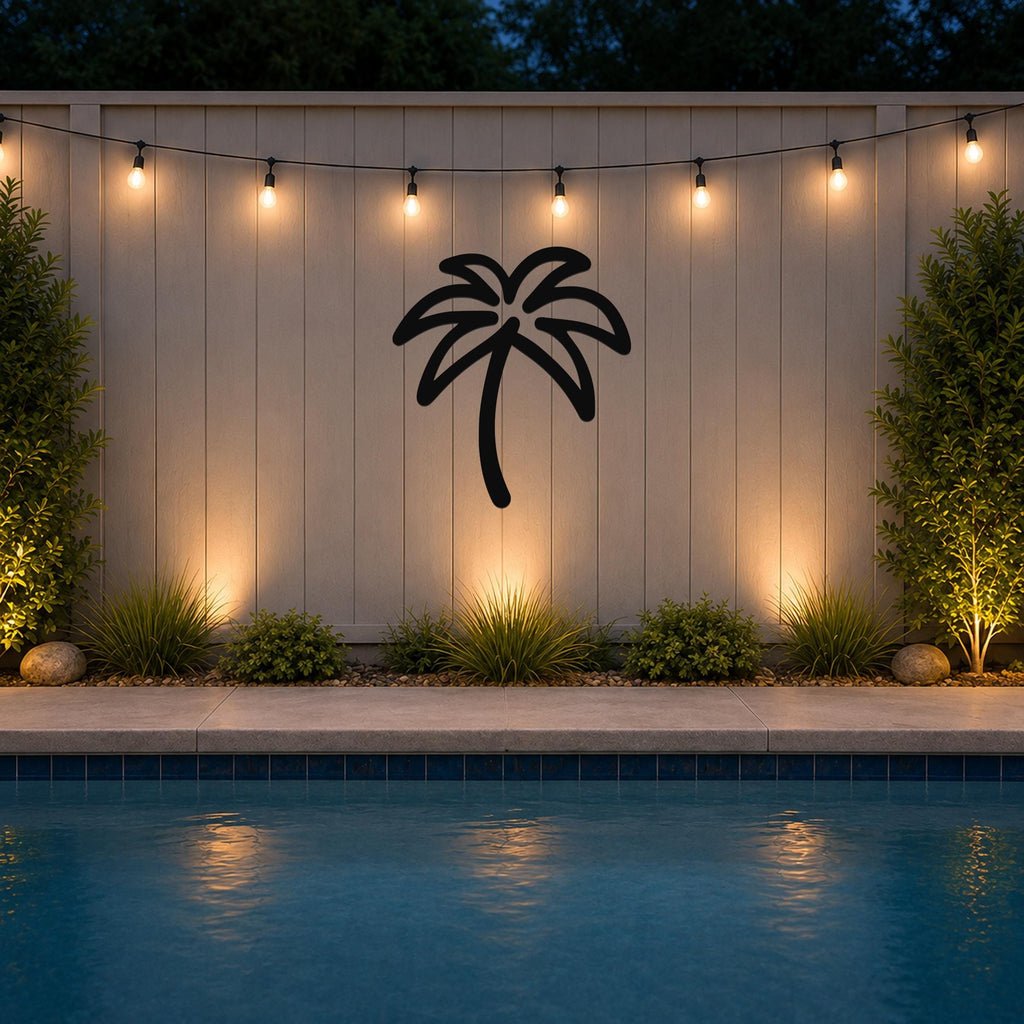 Black metal palm tree wall art with a silhouette design, mounted on a light-colored outdoor wall above a pool, illuminated by string lights.