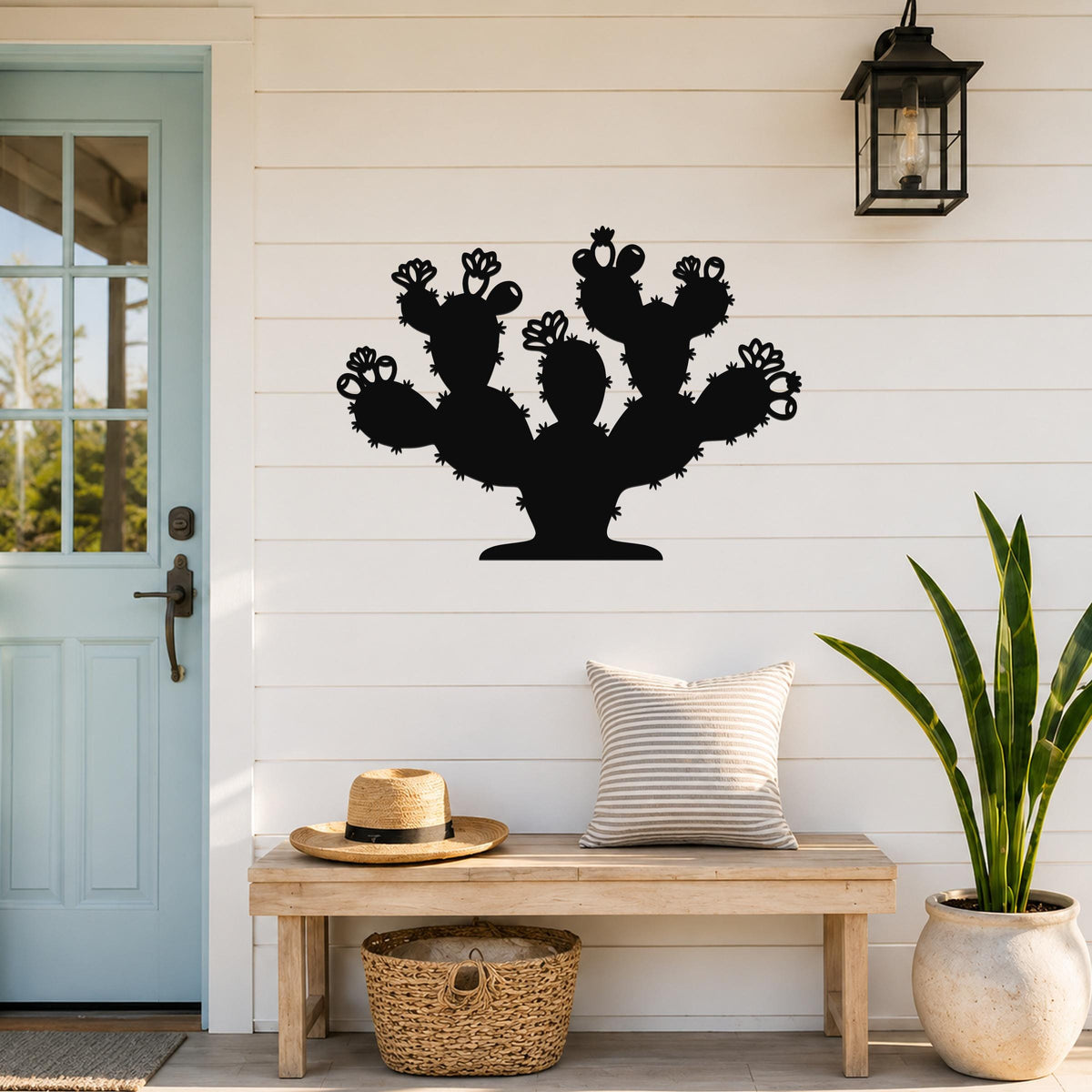 Black metal prickly pear cactus wall art with flower and spine cutouts, mounted on a white shiplap wall above a wooden bench in a porch entryway.