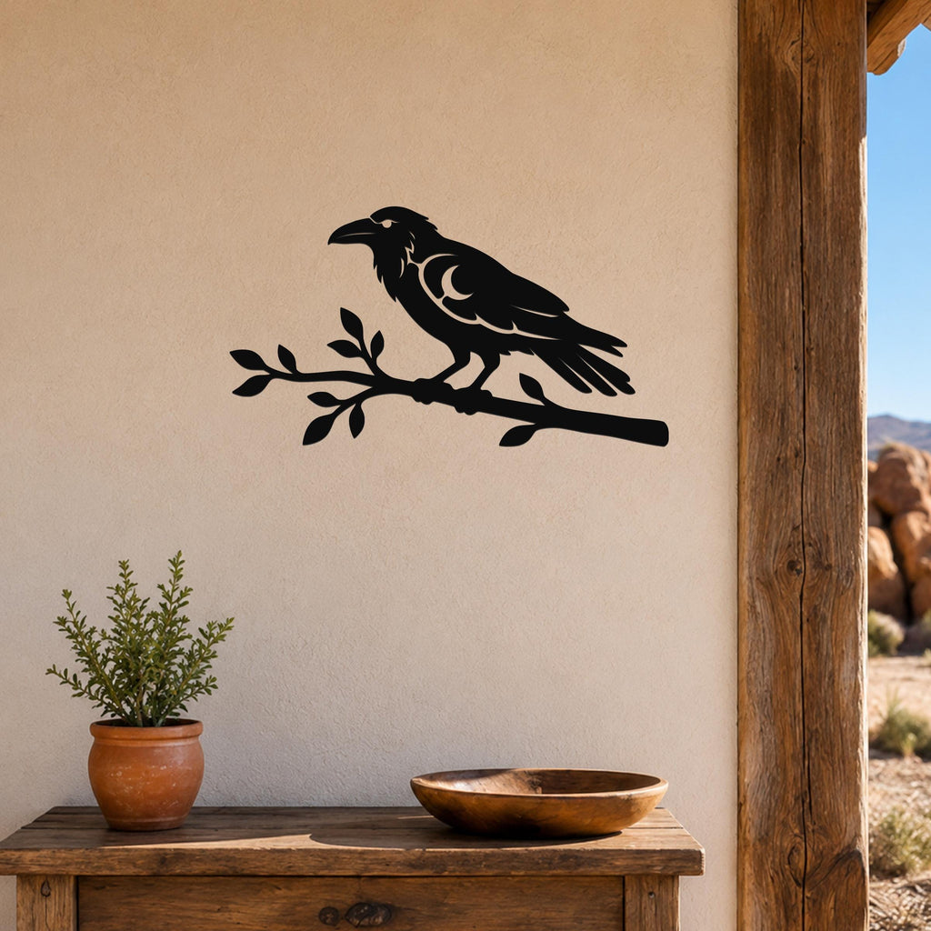Black metal raven wall art cutout perched on a branch, mounted on a beige wall above a wooden table in a rustic outdoor entryway.