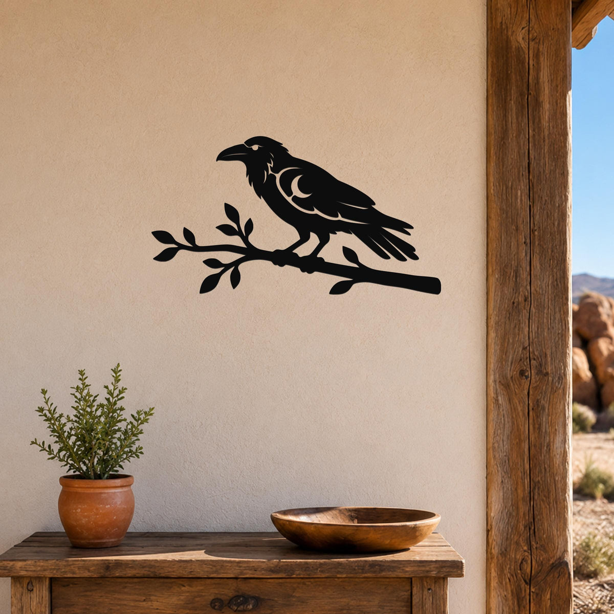 Black metal raven wall art cutout perched on a branch, mounted on a beige wall above a wooden table in a rustic outdoor entryway.