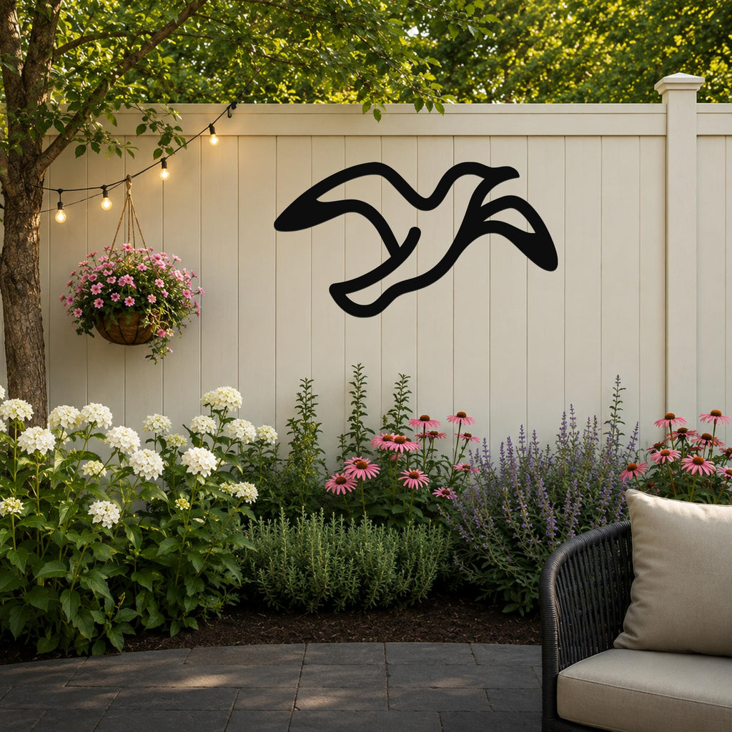Black metal seagull wall art with flowing motion lines, mounted on a white outdoor fence above a garden bed with flowers and string lights.