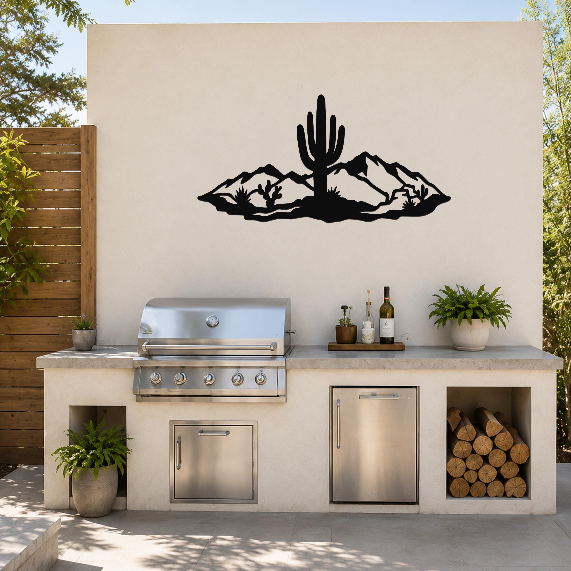 Black metal wall art featuring a desert silhouette with a saguaro cactus and mountains, mounted on an outdoor kitchen wall above a stainless steel grill.