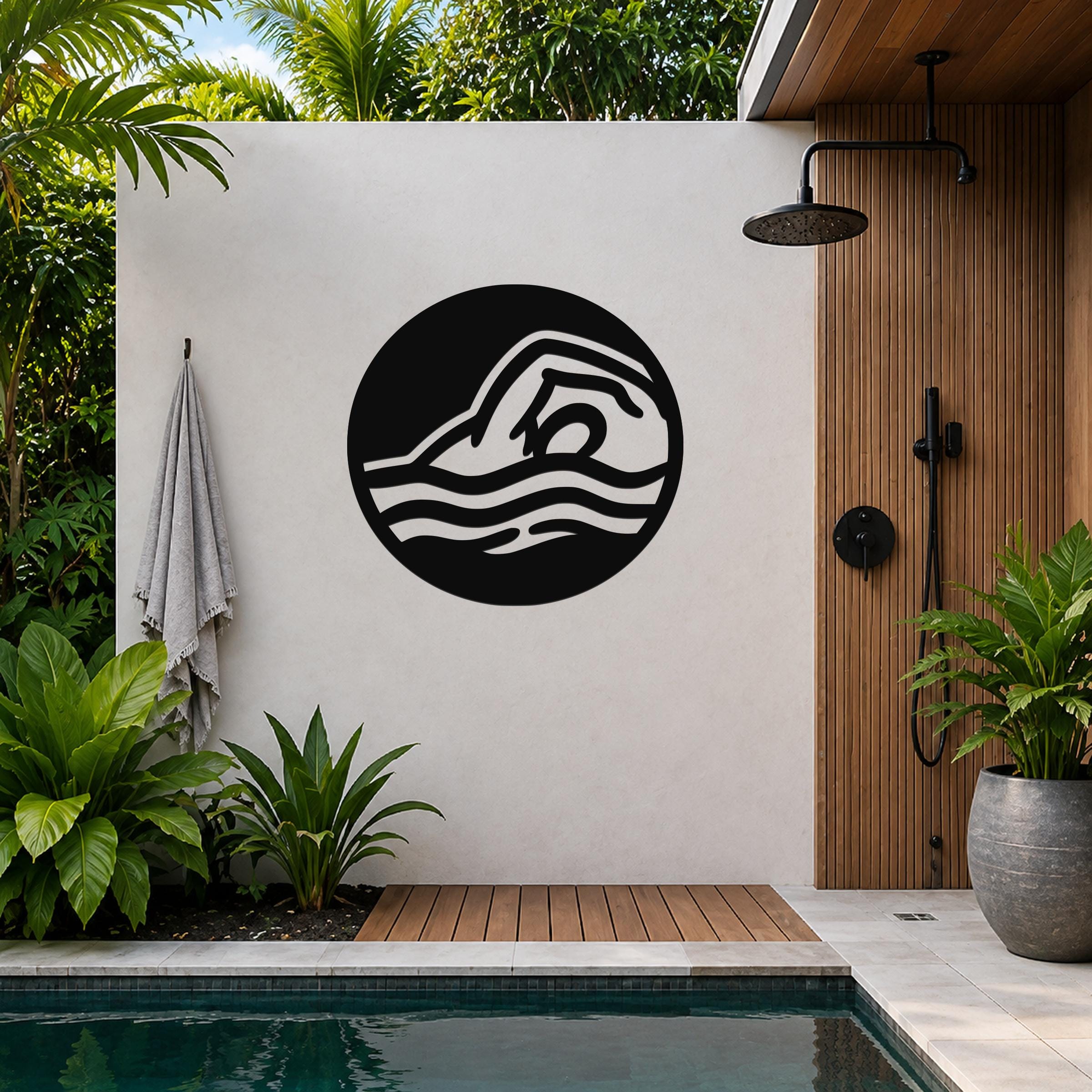 Black metal wall art with a swimmer silhouette and wave cutouts, mounted on a white outdoor wall beside a pool and shower area.