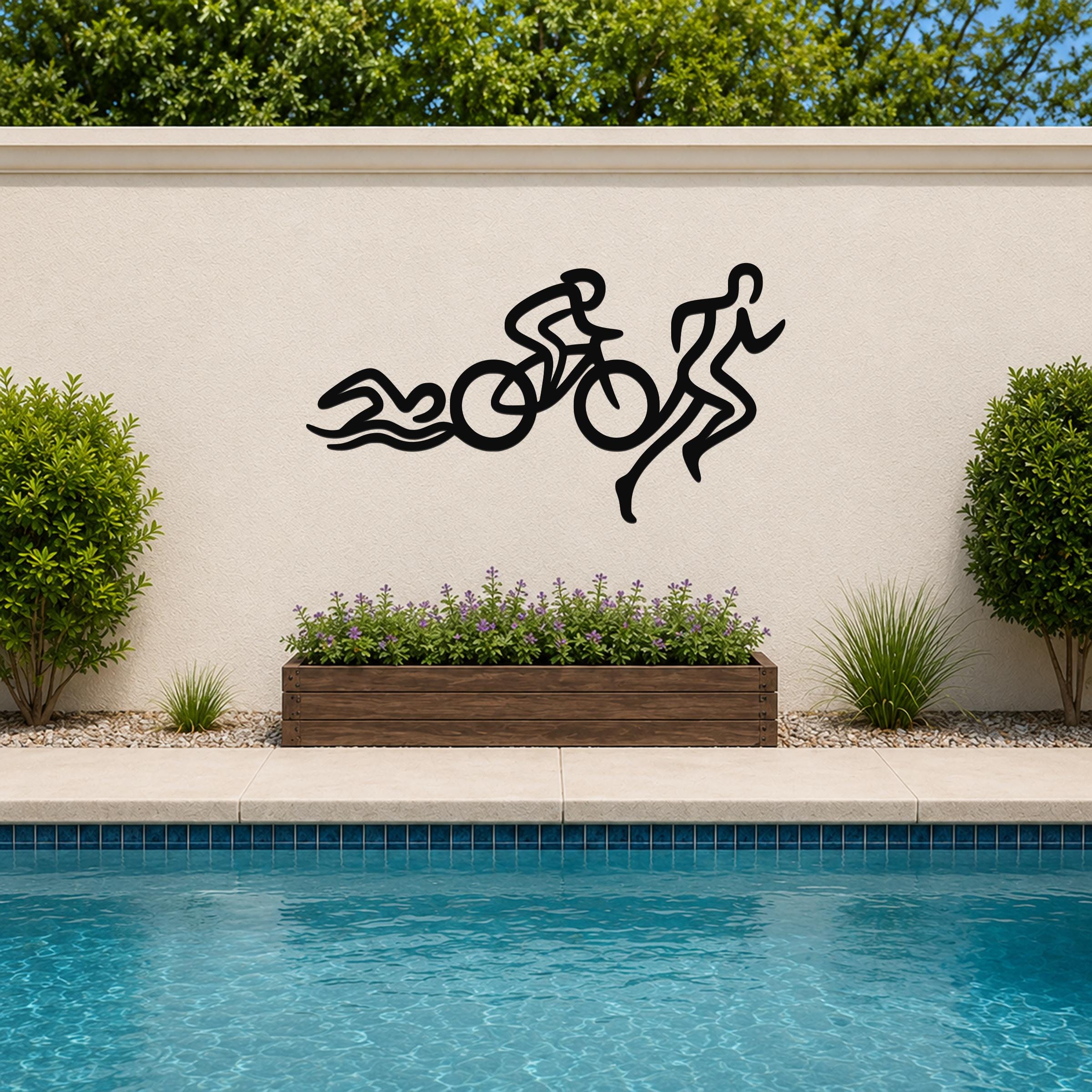 Black metal triathlon wall art with swim, bike, and run silhouette cutouts, mounted on an outdoor wall above a poolside planter.