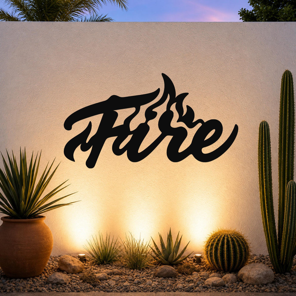 Black metal wall art with the word "Fire" in a fluid, flame-like script, mounted on an outdoor wall above a desert garden with cacti and warm uplighting.