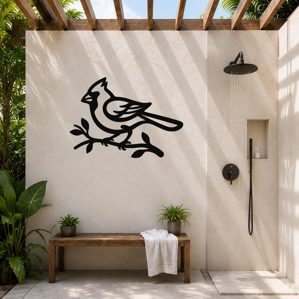 Black metal cardinal wall art in silhouette, perched on a branch, mounted on a beige outdoor shower wall beside a rainfall showerhead.