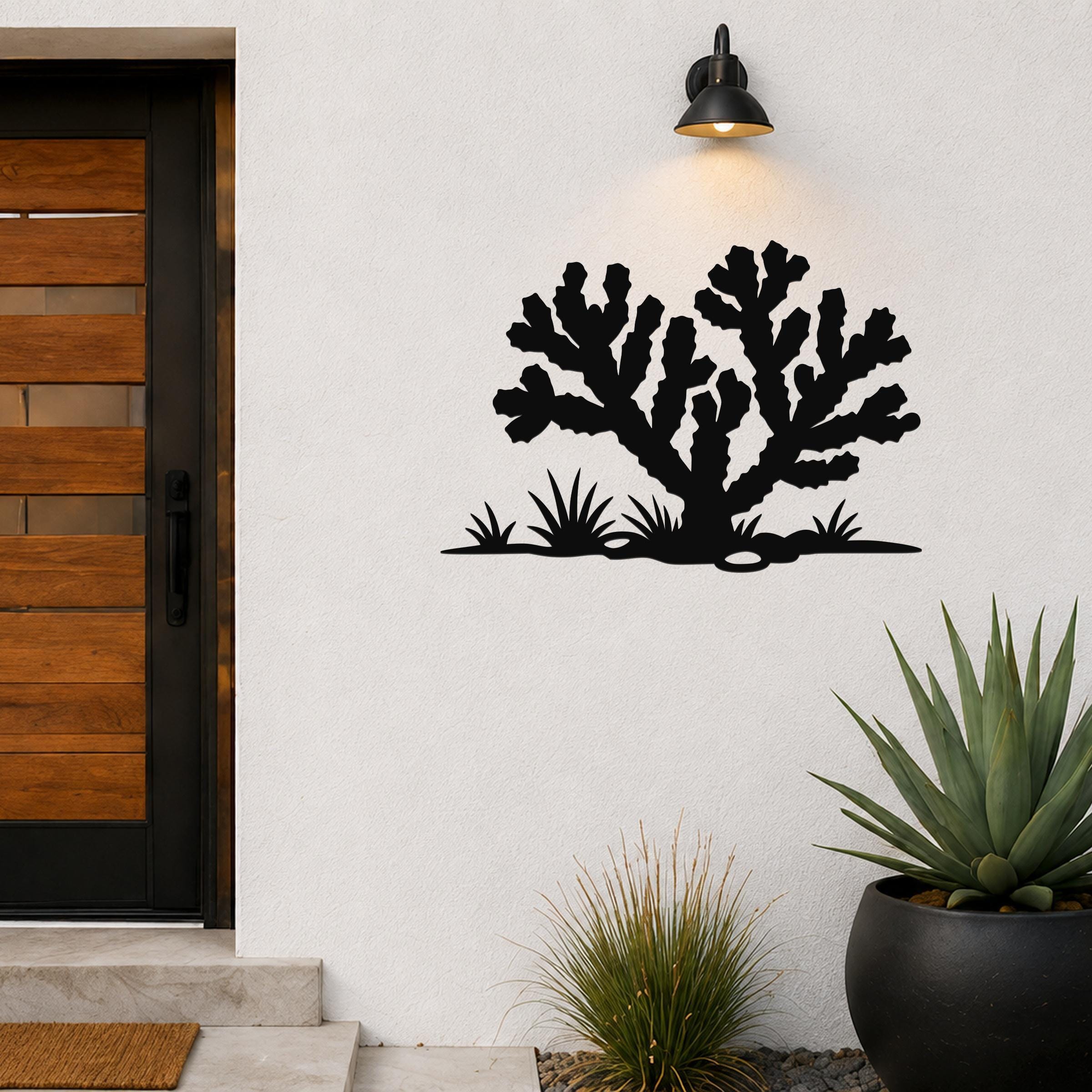 Black metal wall art cutout of a cholla cactus with smaller desert plants, mounted on a white exterior wall beside a wooden door and above potted succulents.