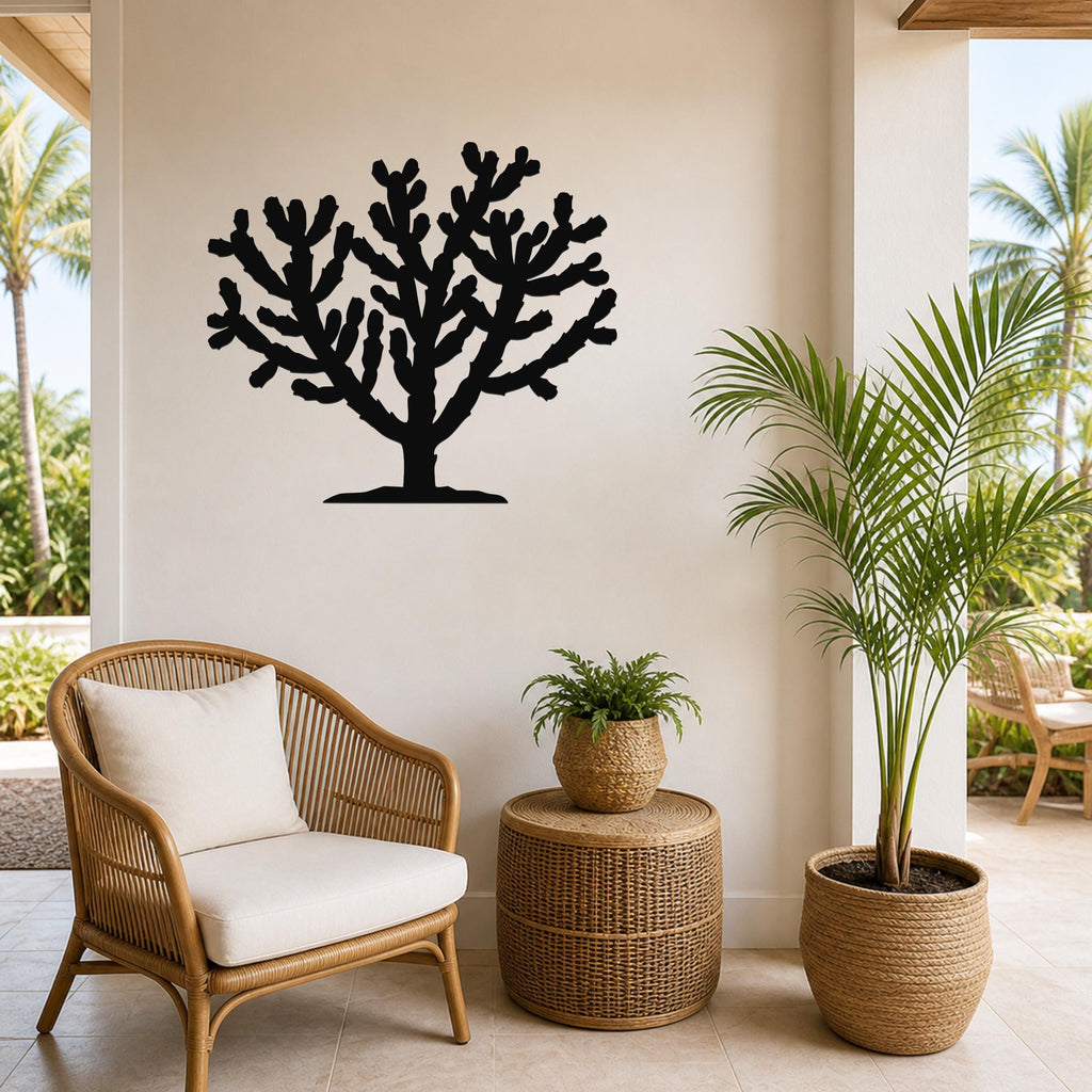 Black metal Cholla cactus wall art silhouette with branching arms, mounted on a light-colored wall above a wicker chair in a tropical patio setting.