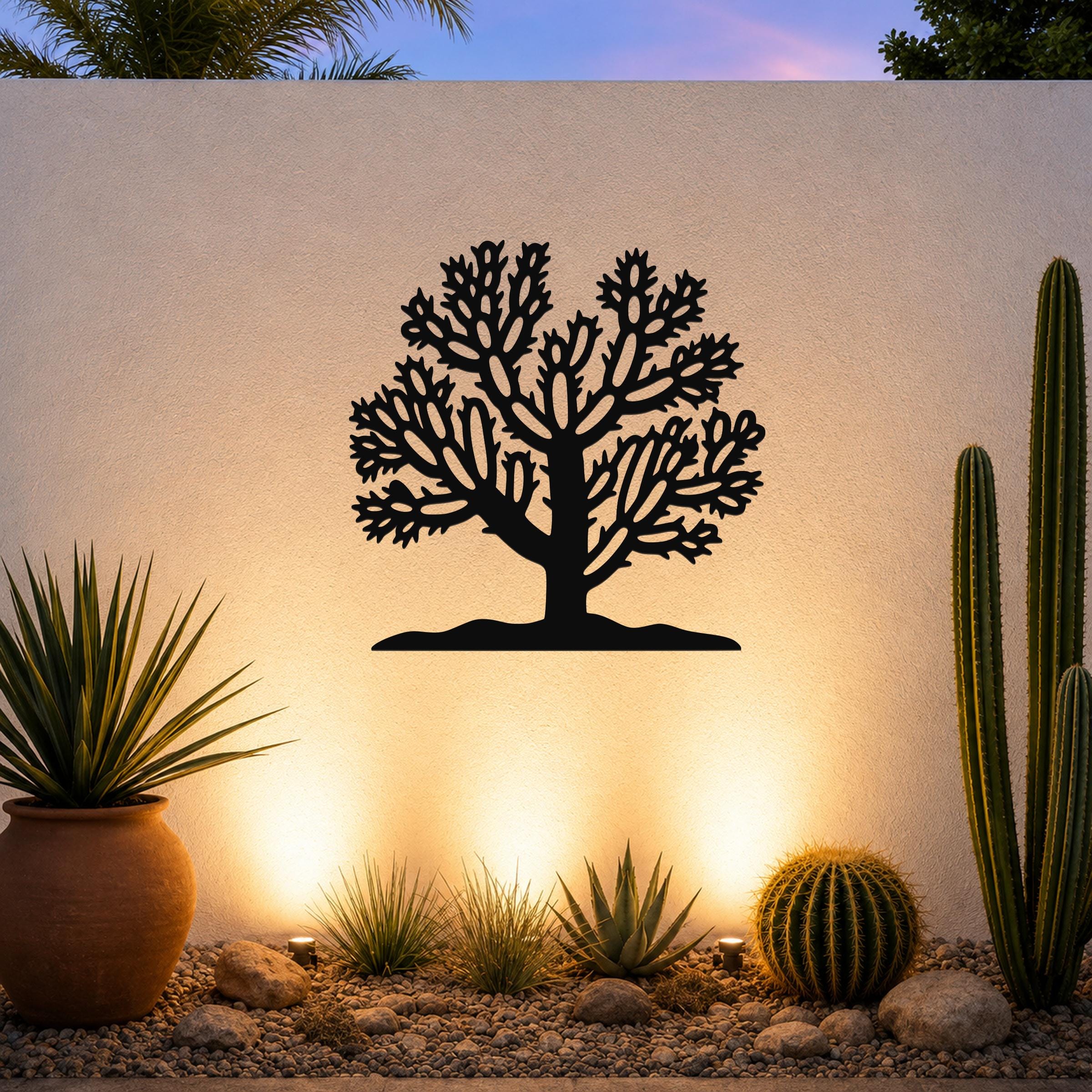 Black metal Cholla cactus wall art silhouette with intricate branch cutouts, mounted on a textured outdoor wall above a desert garden with cacti and warm uplighting.