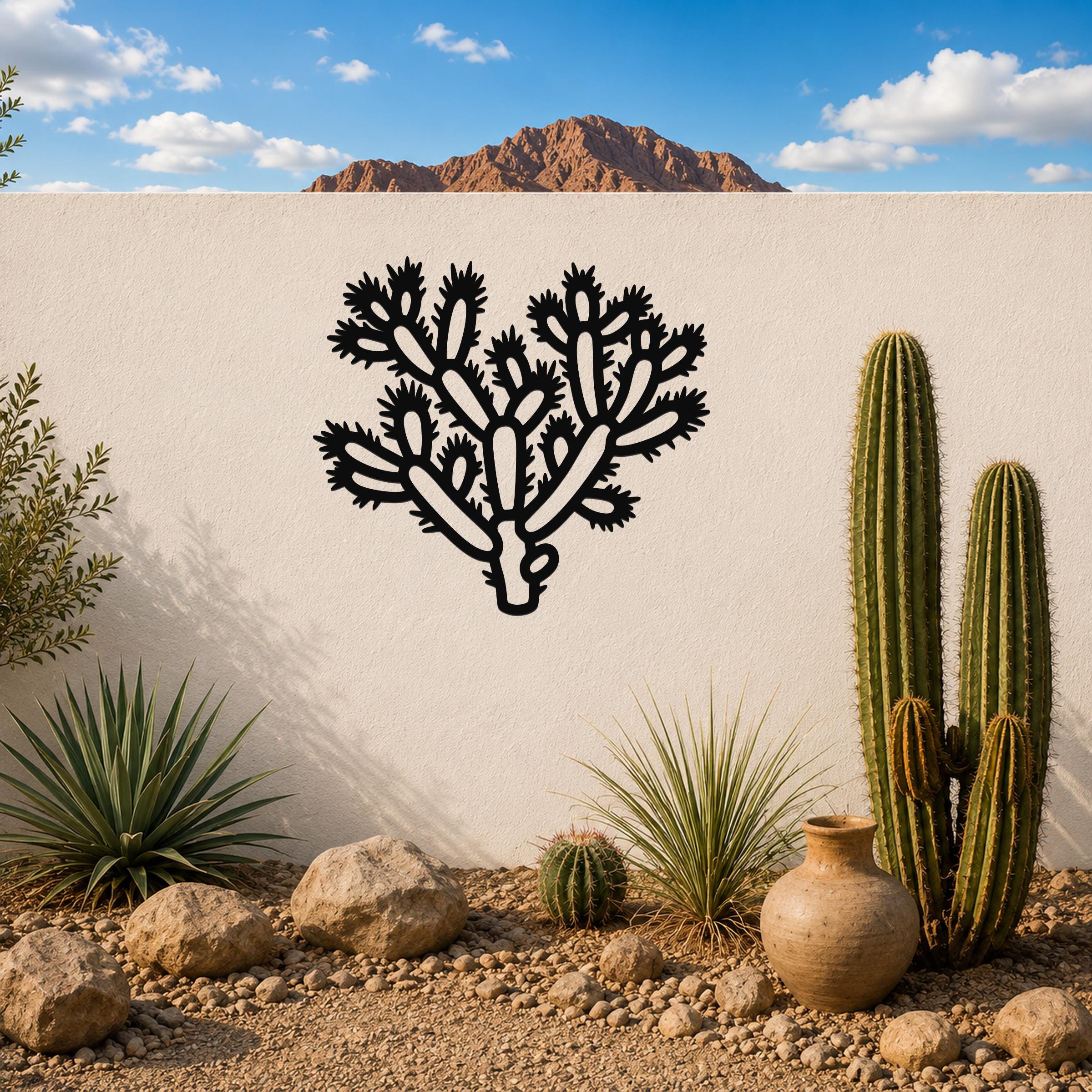 Black metal Cholla cactus wall art with detailed branch and spine cutouts, mounted on a white outdoor wall above a desert garden with real cacti and rocks.