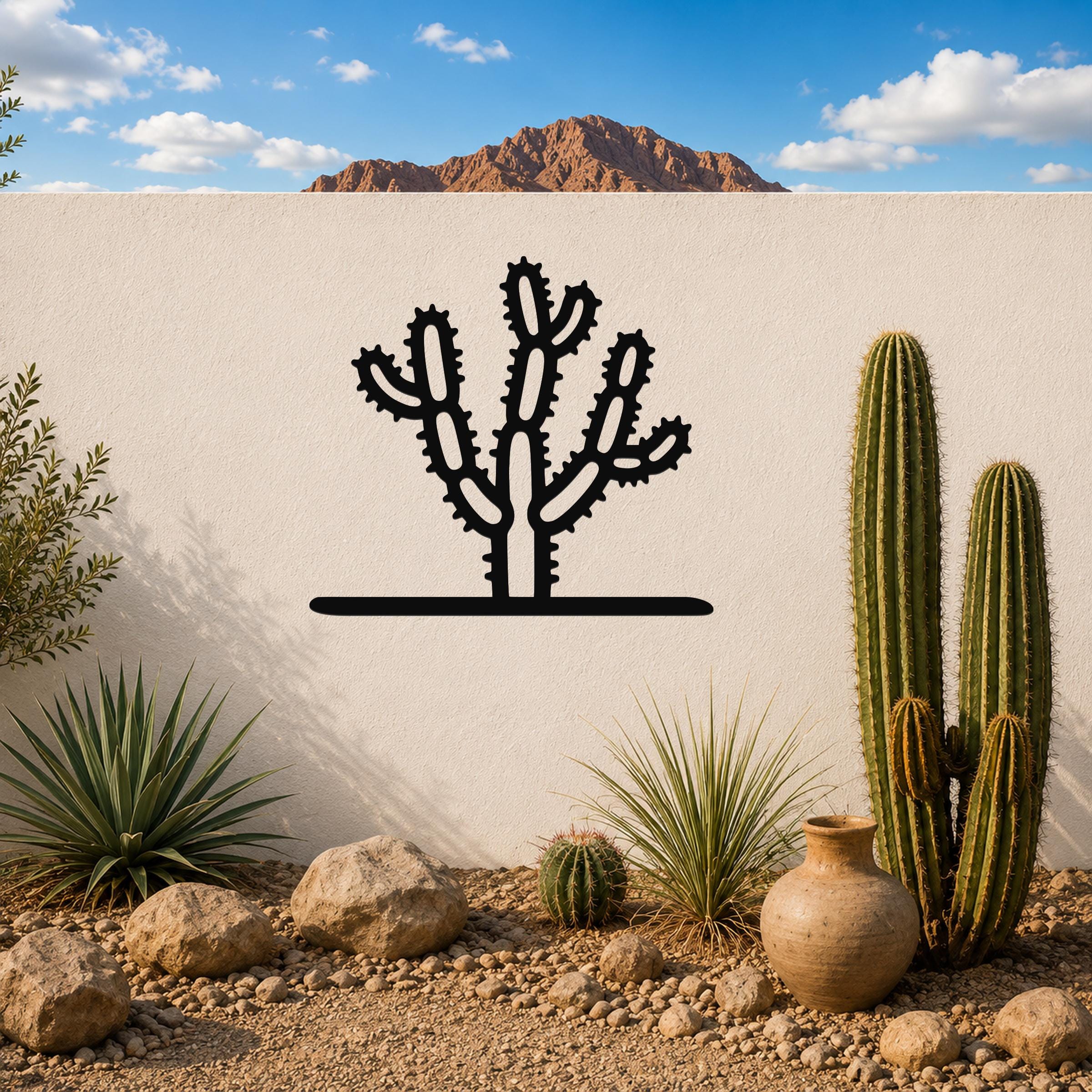 Black metal cholla cactus wall art with spiny branch cutouts, mounted on a white outdoor wall above a desert garden with real cacti and rocks.