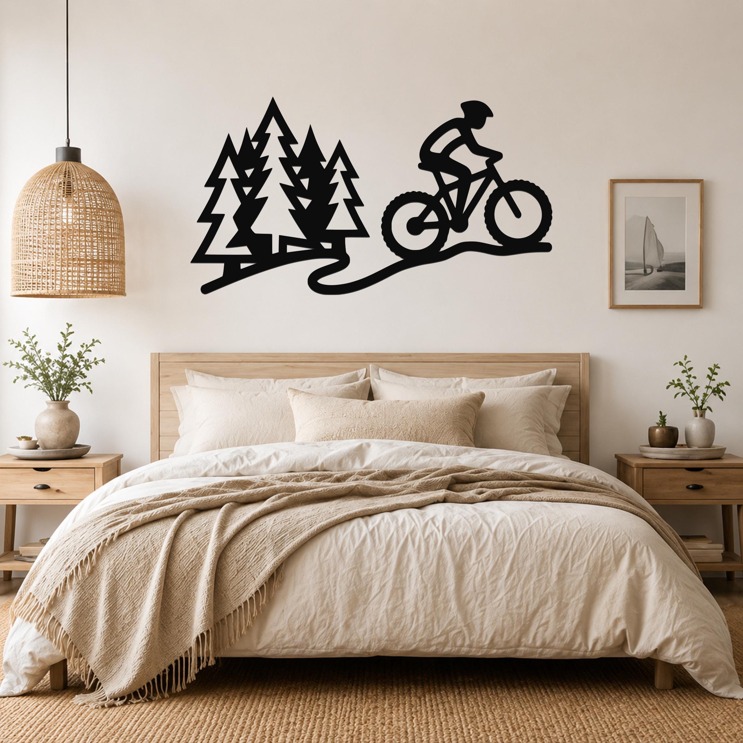 Black metal wall art silhouette of a cyclist on a forest trail with pine trees, mounted above a bed in a bedroom.