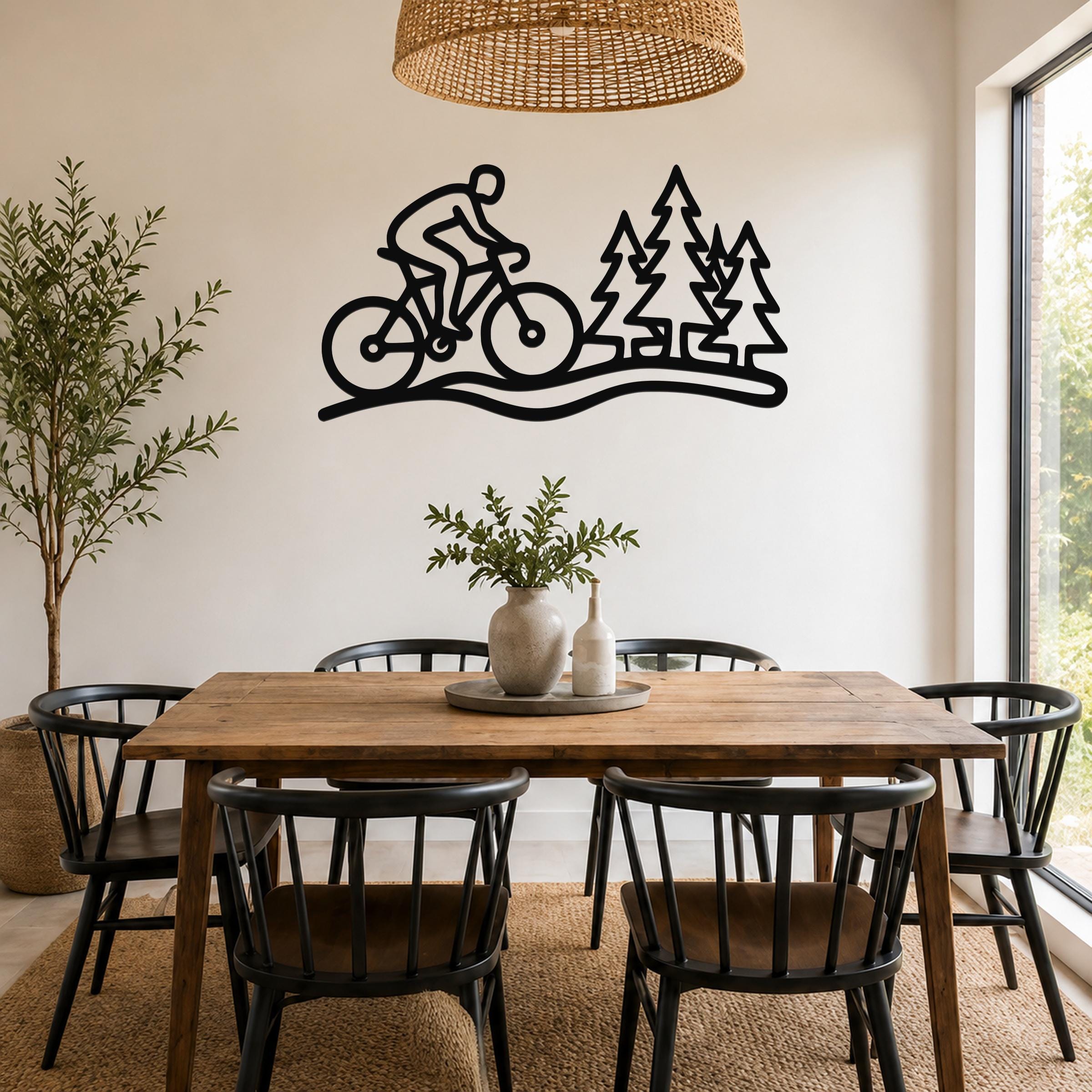 Black metal wall art silhouette of a cyclist on a trail with pine trees, mounted above a wooden dining table in a bright room.