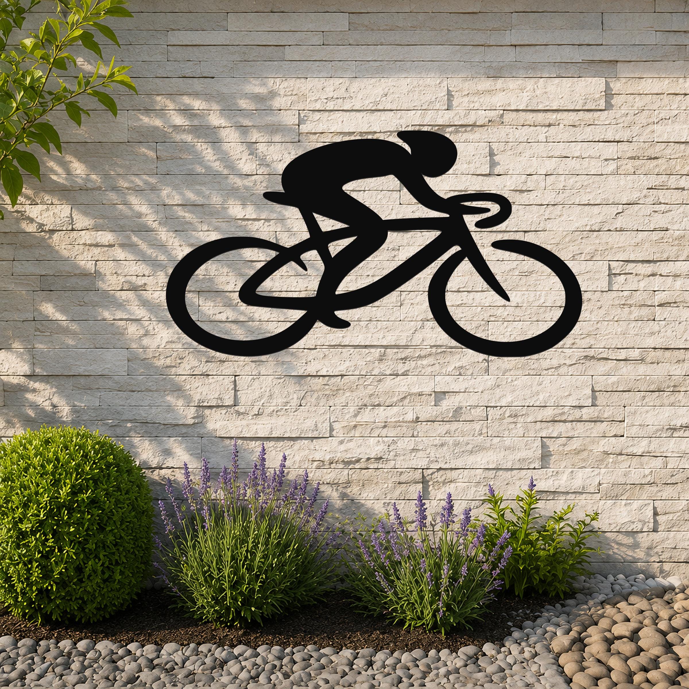 Black metal wall art in a bold silhouette of a cyclist on a bike, mounted on a light stone wall above a garden bed with lavender and pebbles.