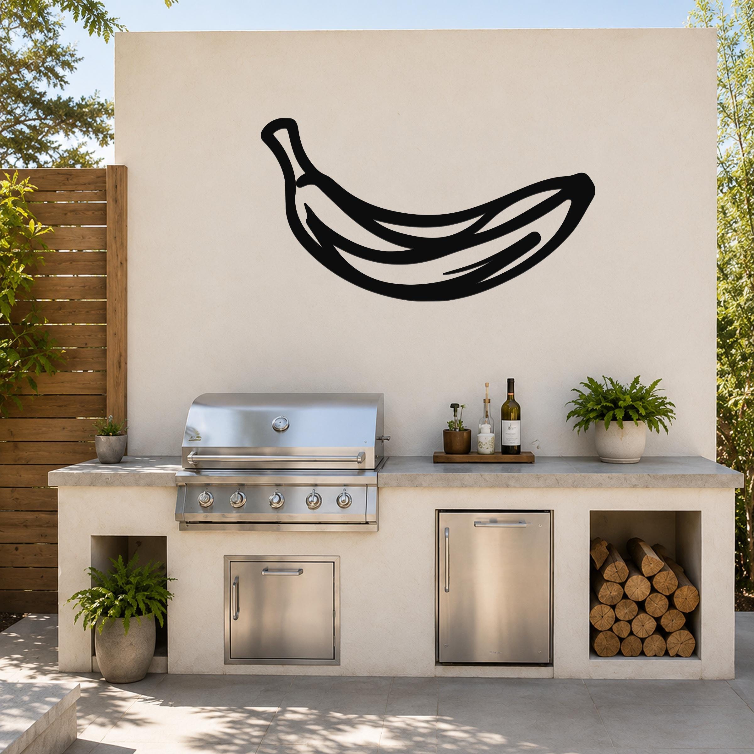 Black metal banana wall art silhouette mounted on an outdoor kitchen wall above a stainless steel grill and counter.