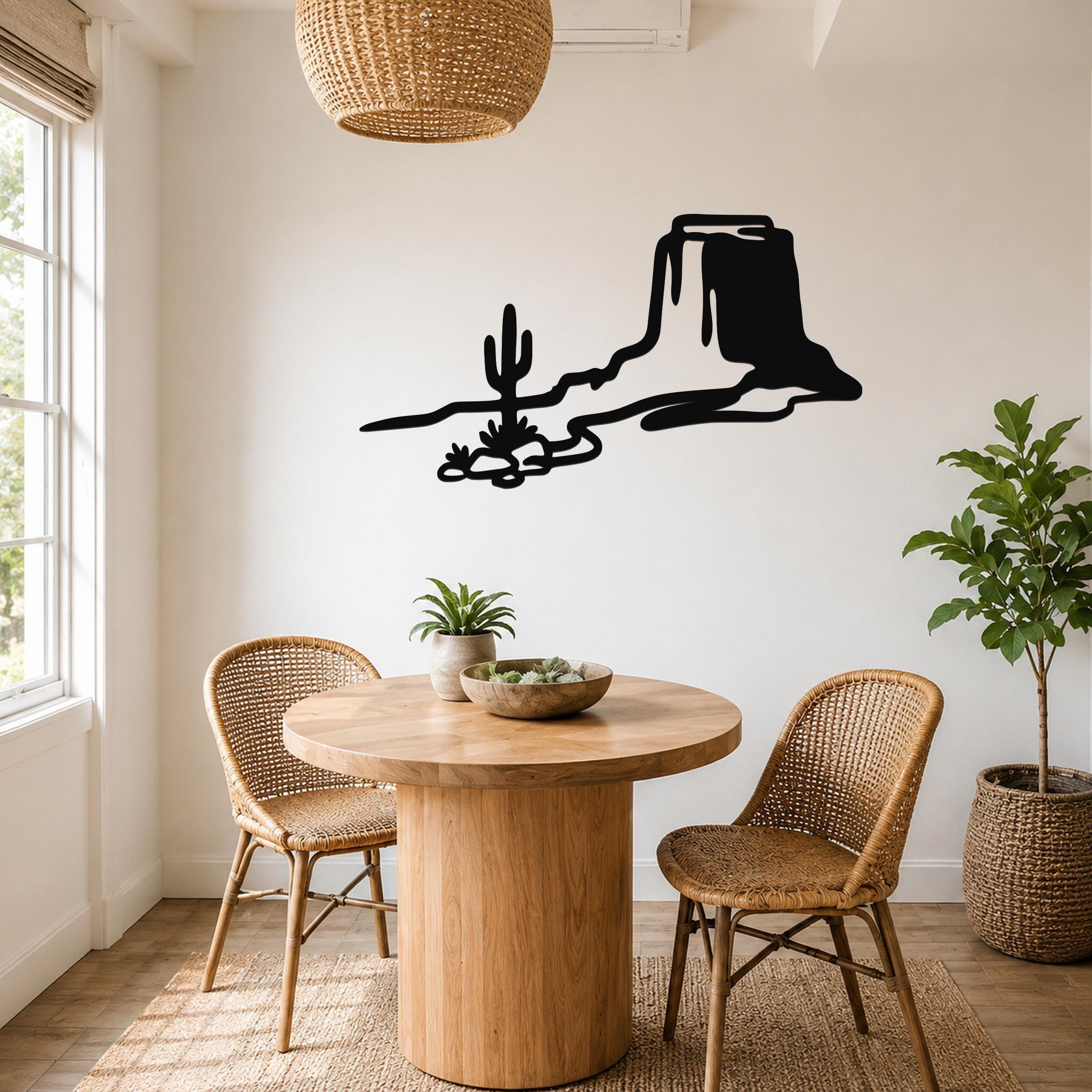 Black metal wall art silhouette of a desert mesa and cactus, mounted on a white wall above a round wooden table in a bright dining nook.