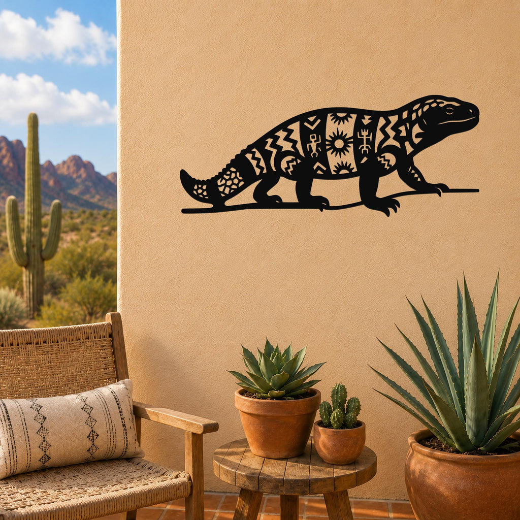 Black metal wall art of a Gila monster with intricate geometric cutouts, mounted on a tan outdoor wall above a small wooden table with potted succulents.