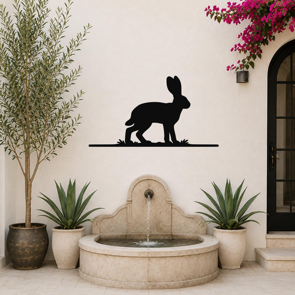Black metal jackrabbit wall art silhouette mounted on a white outdoor wall above a stone fountain, flanked by potted plants and a blooming vine.