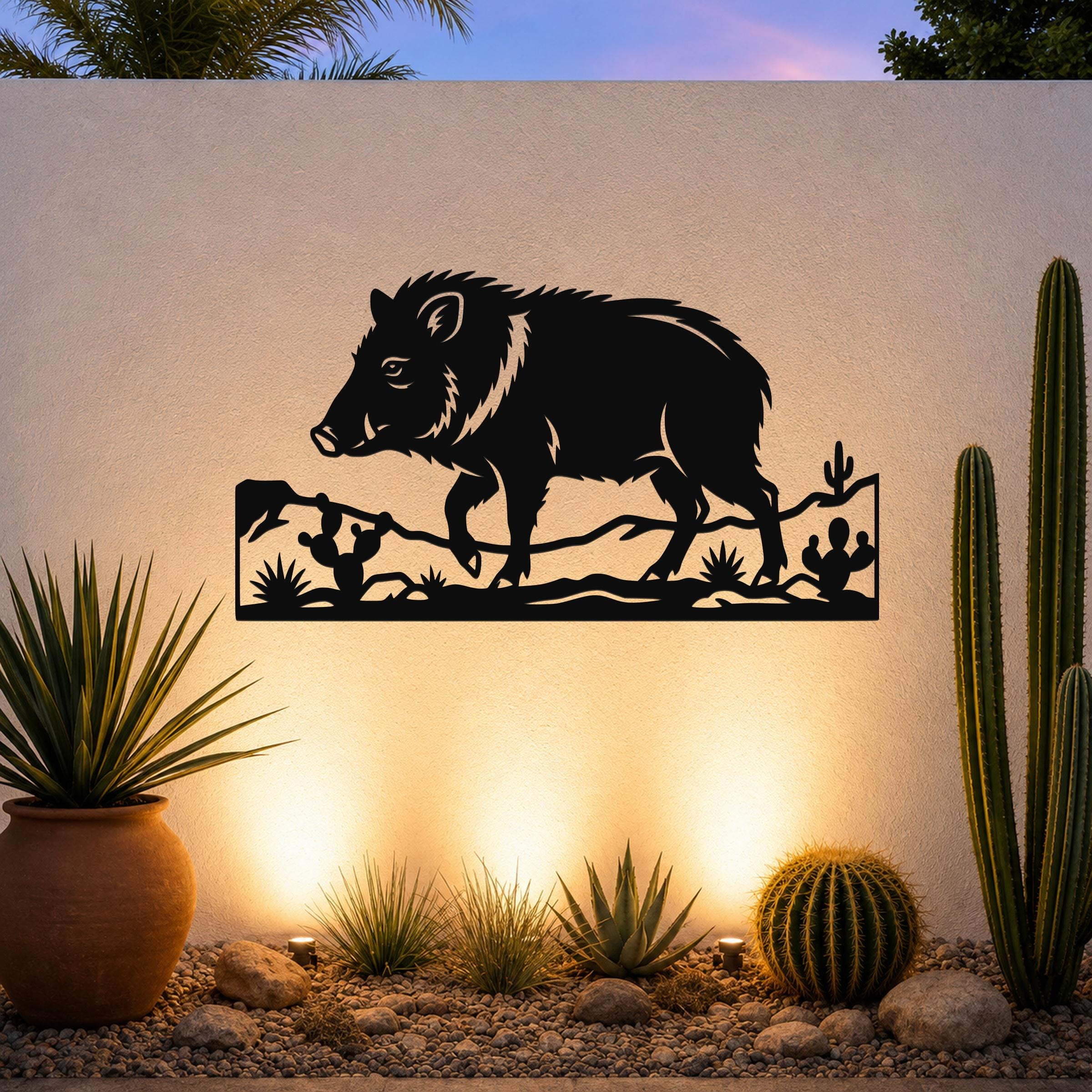 Black metal wall art featuring a javelina silhouette with cacti and desert terrain cutouts, mounted on an outdoor wall above a gravel garden with potted succulents.