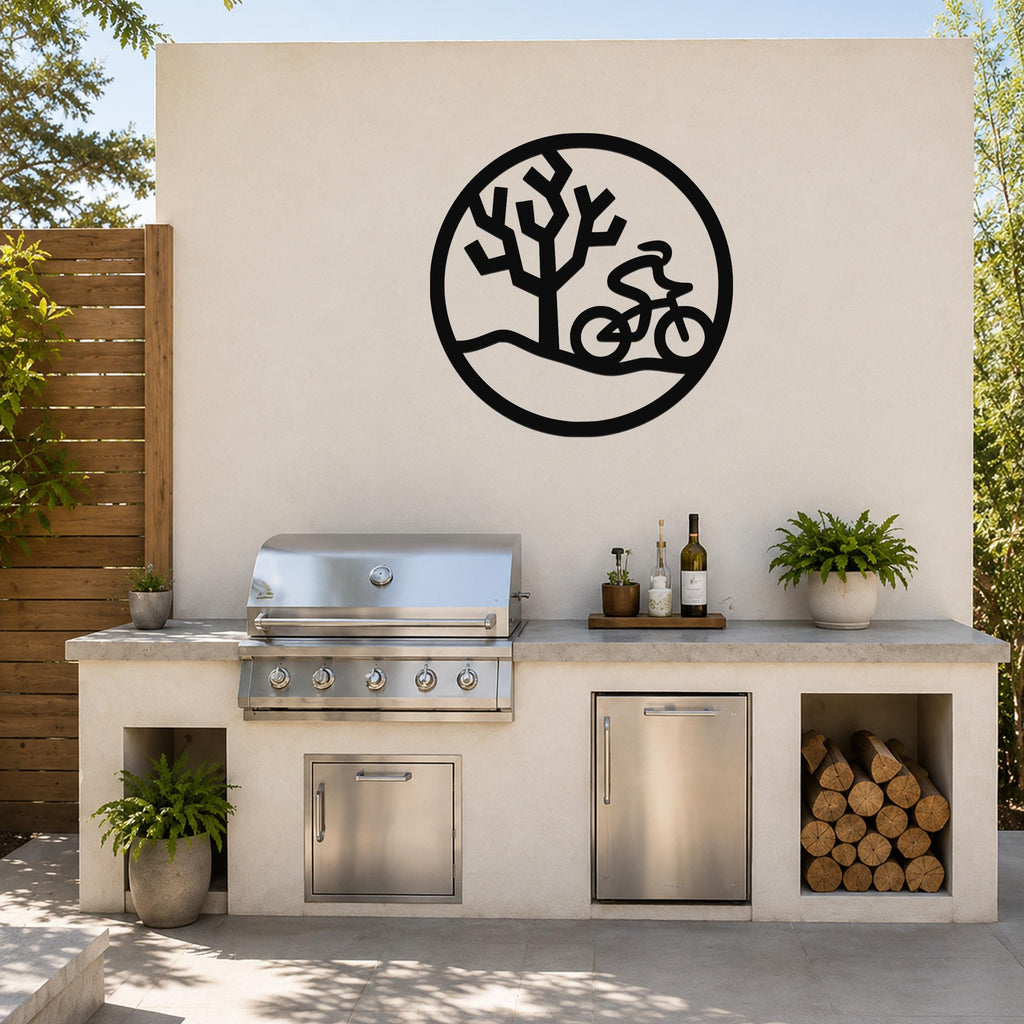 Black metal wall art with a circular silhouette of a cyclist and a Joshua tree, mounted on an outdoor kitchen wall above a stainless steel grill.